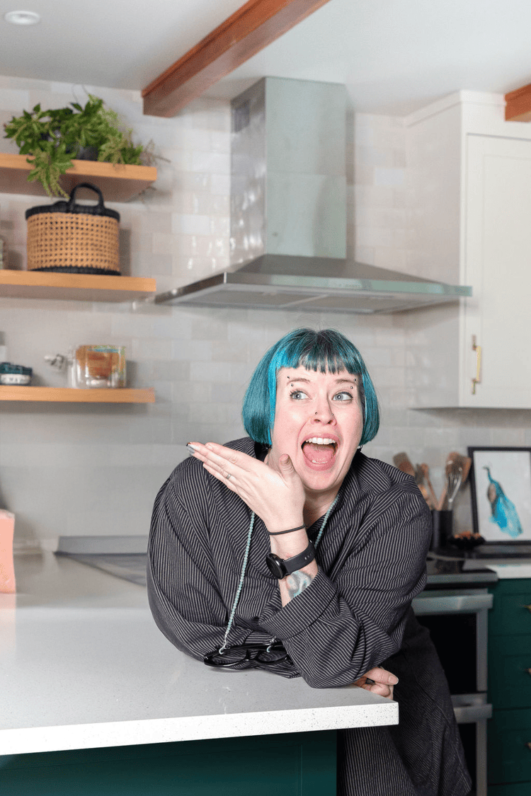 Woman with teal hair making a surprised expression, leaning on a kitchen counter in a modern kitchen with white cabinets and open wooden shelves.