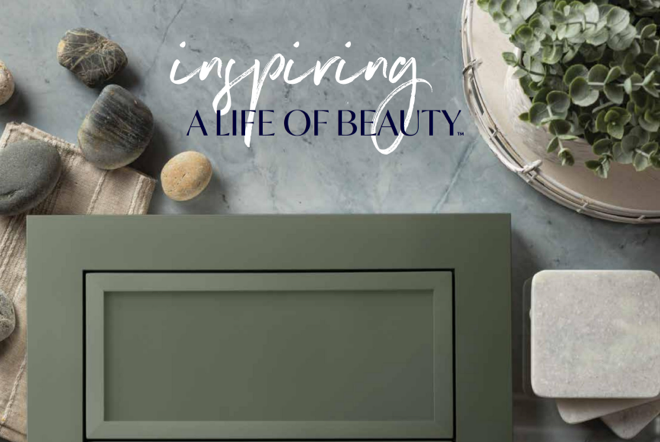 A flat lay of beauty and spa items including rocks, a towel, a box with a scrollwork pattern, a book with the phrase 'inspiring a life of beauty,' a gray box, and a potted plant, on a gray surface.