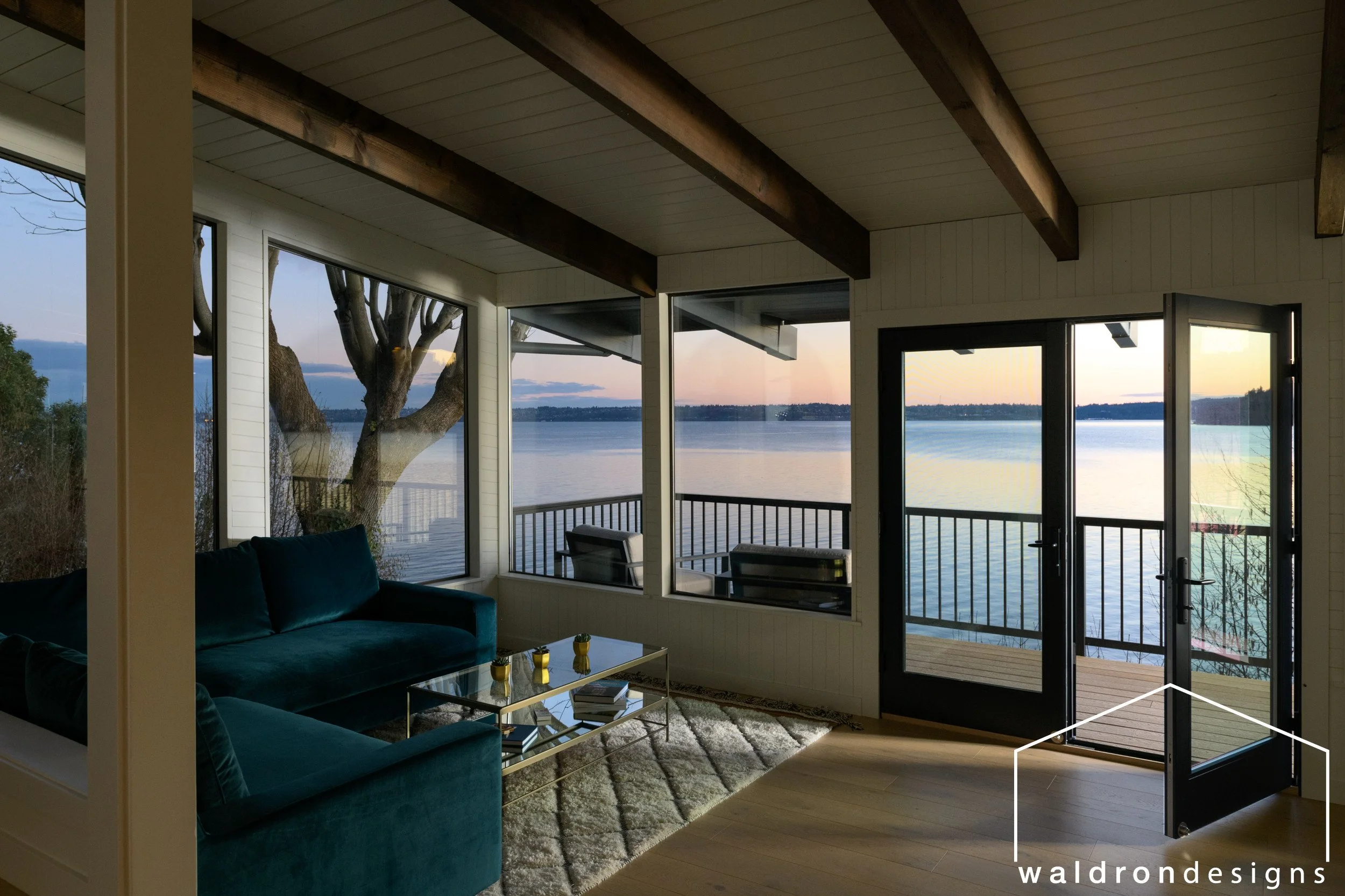 Living room with large windows overlooking a lake, a teal sofa, a glass coffee table, a rug, and an outdoor deck with a railing and lake view during sunset.
