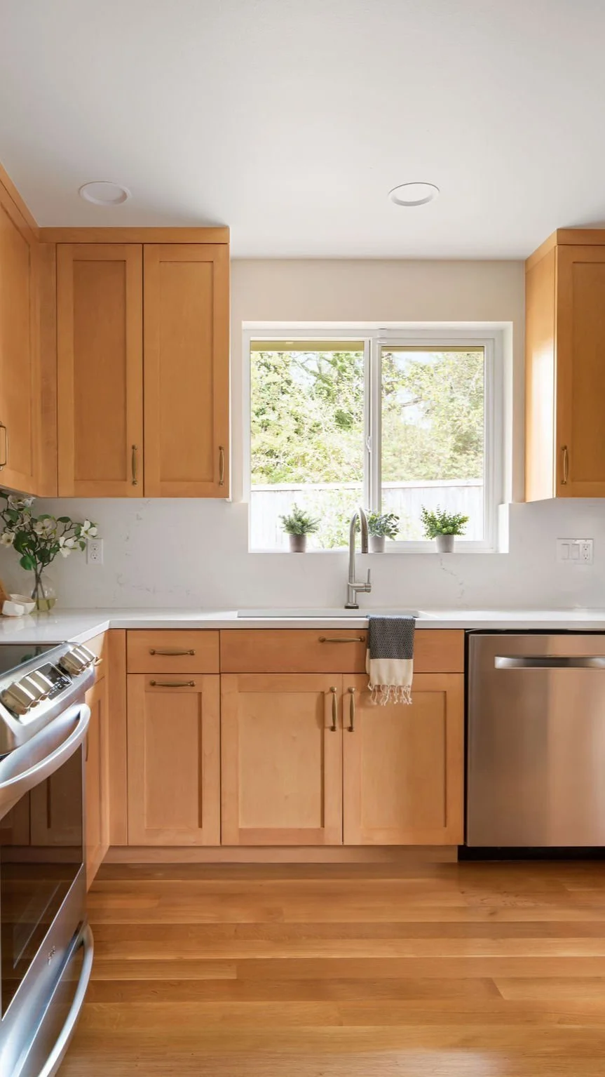 Kitchen with light wood cabinets, stainless steel dishwasher, oven, and a window with three potted plants. White countertop and a white wall, with a small towel hanging from the sink.