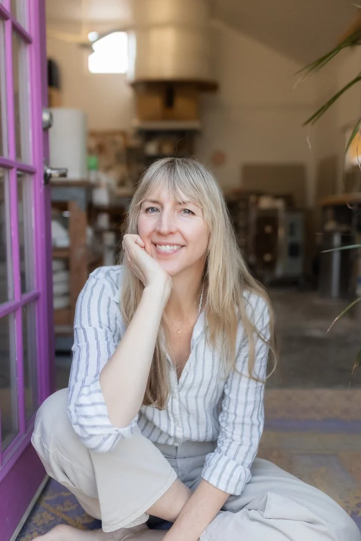 A woman with long blonde hair, wearing a striped button-up shirt, smiling and sitting on the floor in a cozy, well-lit indoor space with a purple door on the left side.