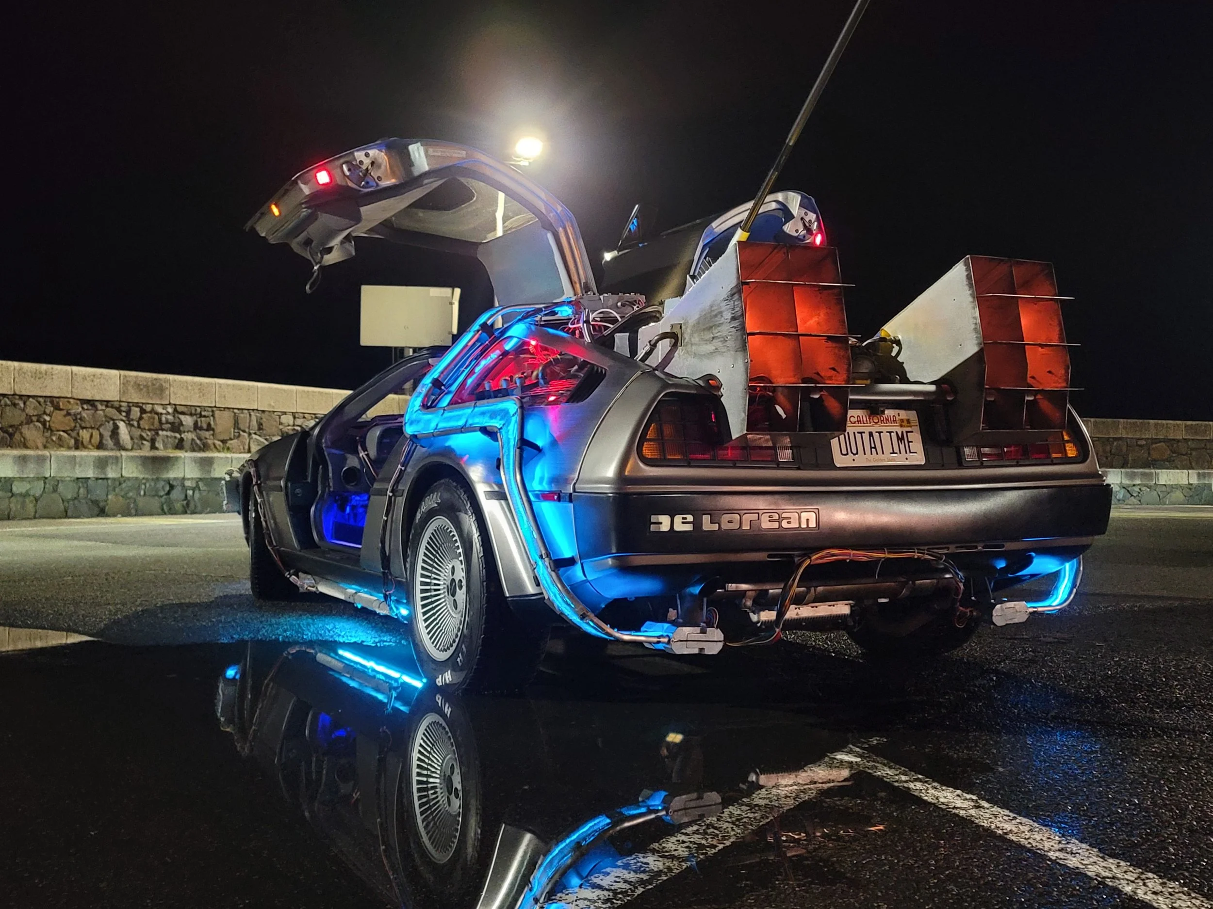 A DeLorean car modified with futuristic components, illuminated with blue lights, parked at night with a reflection on the wet surface underneath. The car has a sign that says 'OUTATIME' on the rear, resembling the time machine from the movie 'Back to the Future,' with exposed wiring and a rear extension resembling a rocket or flux capacitor.