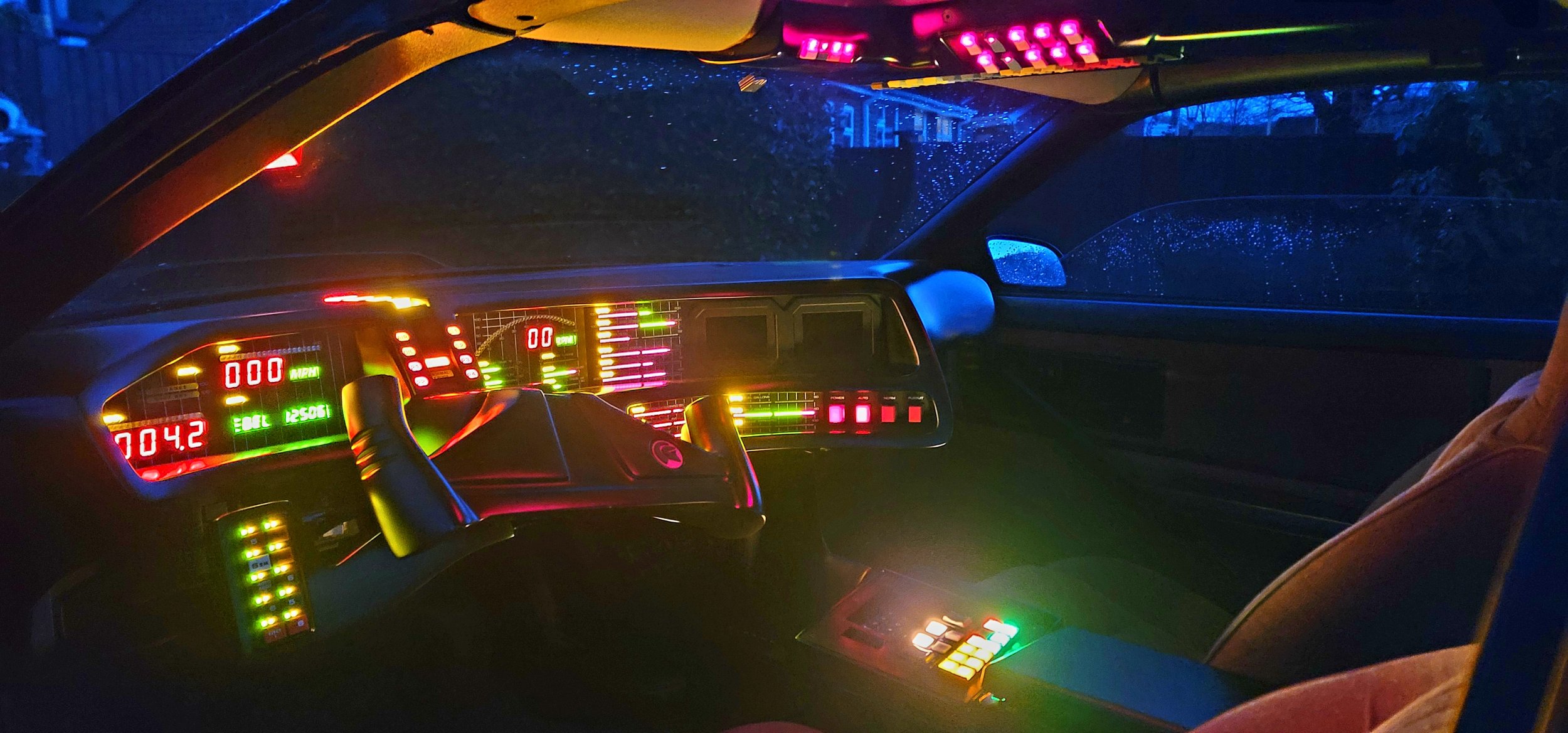 Inside a car at night with illuminated dashboard, dashboard lights in red, green, and pink, and rain droplets on the windshield visible in the background.