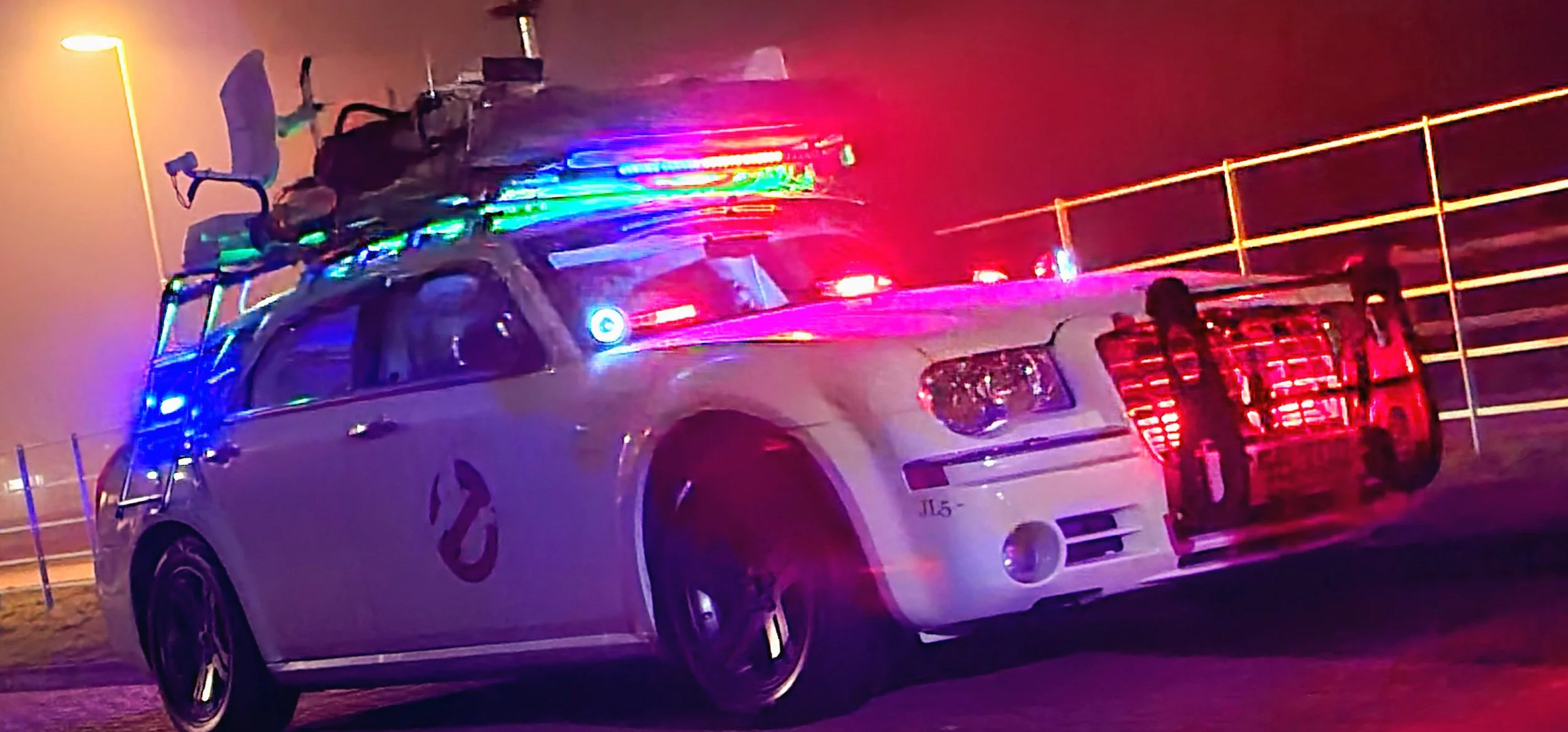 A white Ghostbusters-themed vehicle with colorful lights on the roof, parked near a fence at night, with emergency lights flashing.