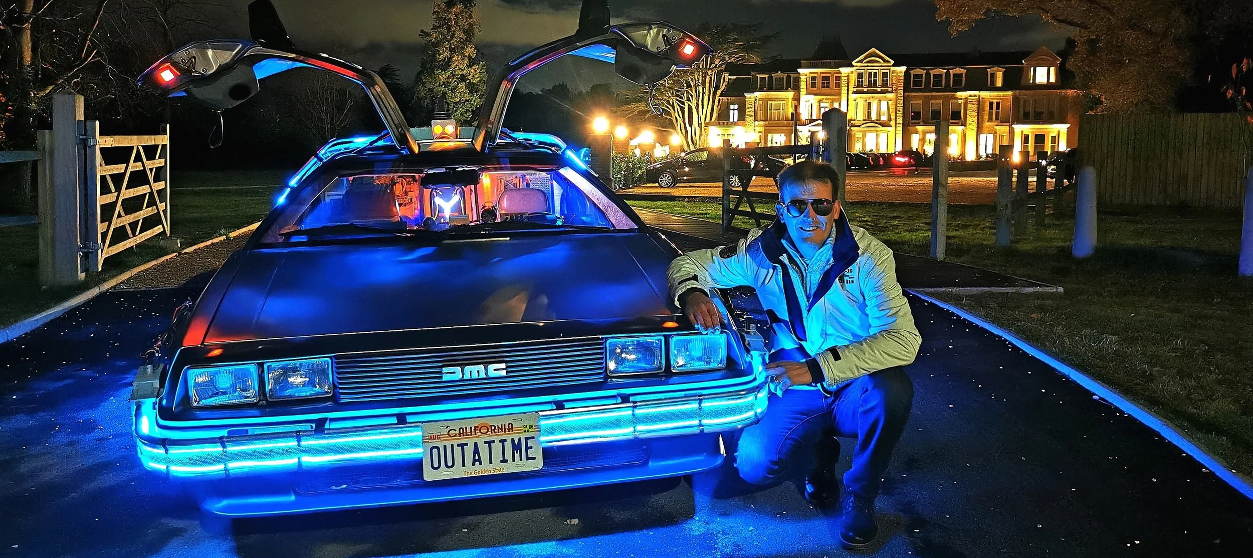 A man wearing sunglasses and a light-colored jacket kneeling next to a DeLorean car with blue neon lights and gull-wing doors. The car has a California license plate that reads OUTATIME. The scene is set at night with a well-lit house and parked cars