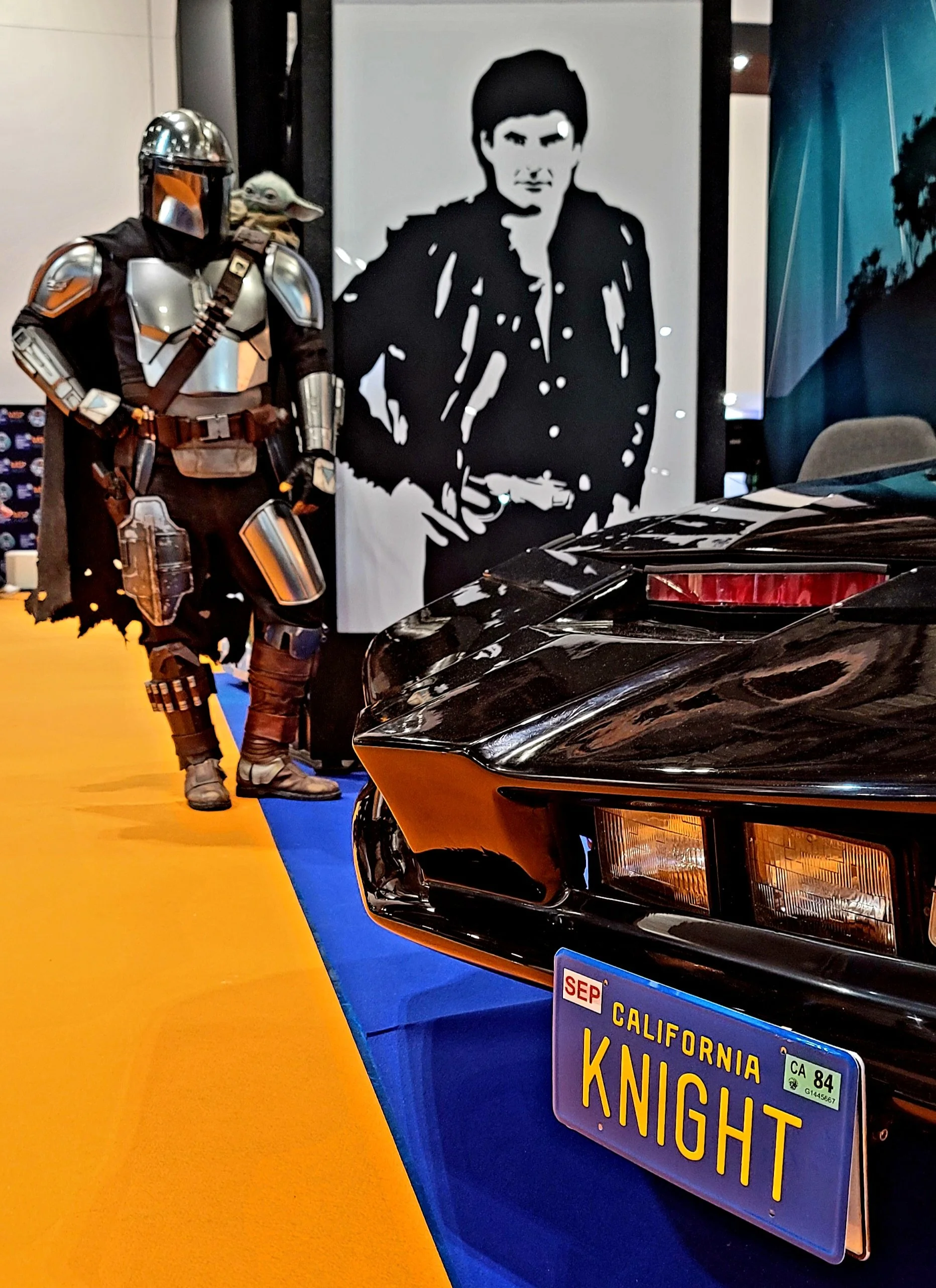 A black sports car with a California license plate that reads 'KNIGHT' and a yellow stripe along the side, parked on a blue and yellow floor at an indoor exhibit. In the background, there is a life-sized statue of Boba Fett from Star Wars and a large black-and-white portrait of a young man with dark hair and a serious expression.