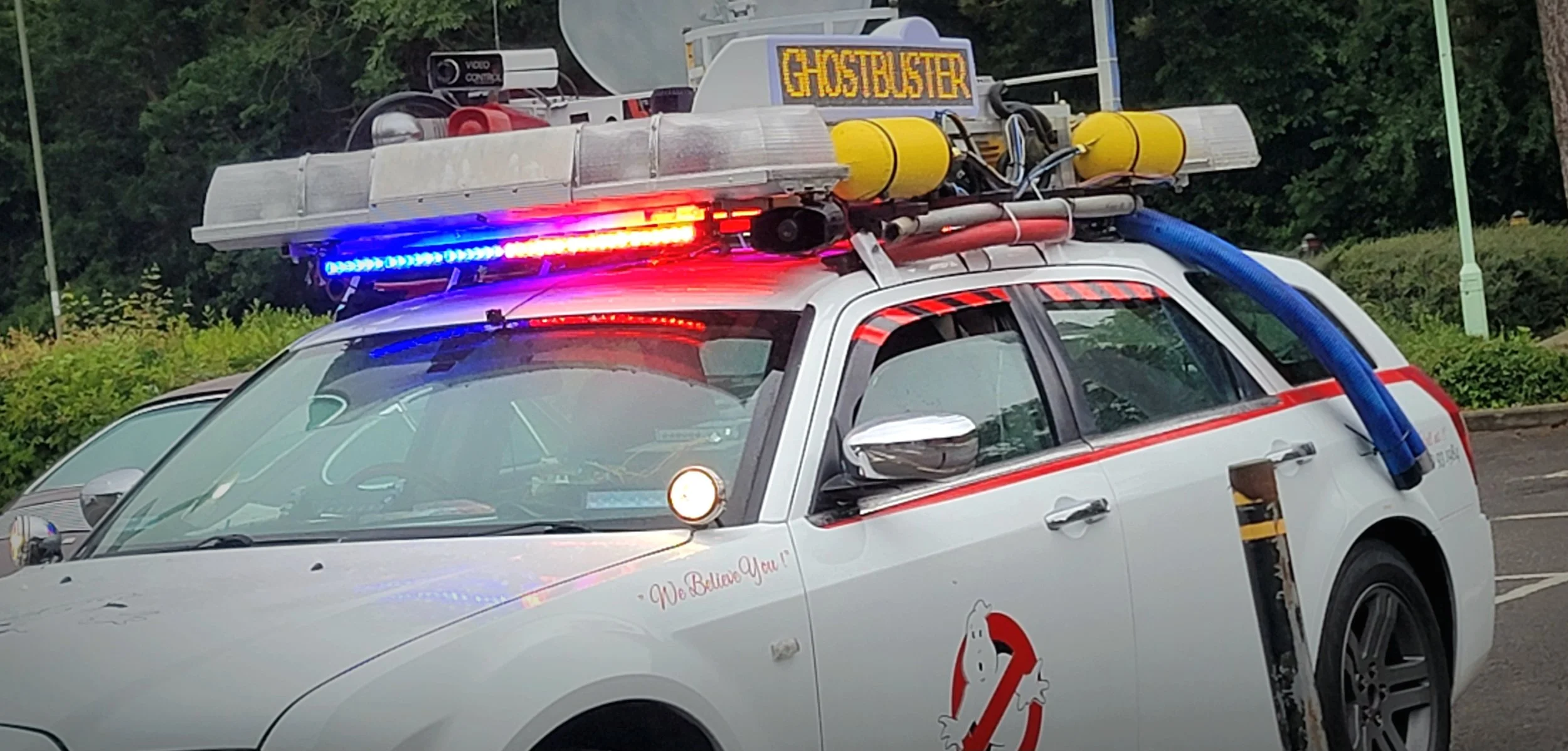 A white vehicle with red and blue flashing lights, decorated with a Ghostbusters theme and logo, equipped with ghost capture equipment on the roof, in a parking lot.