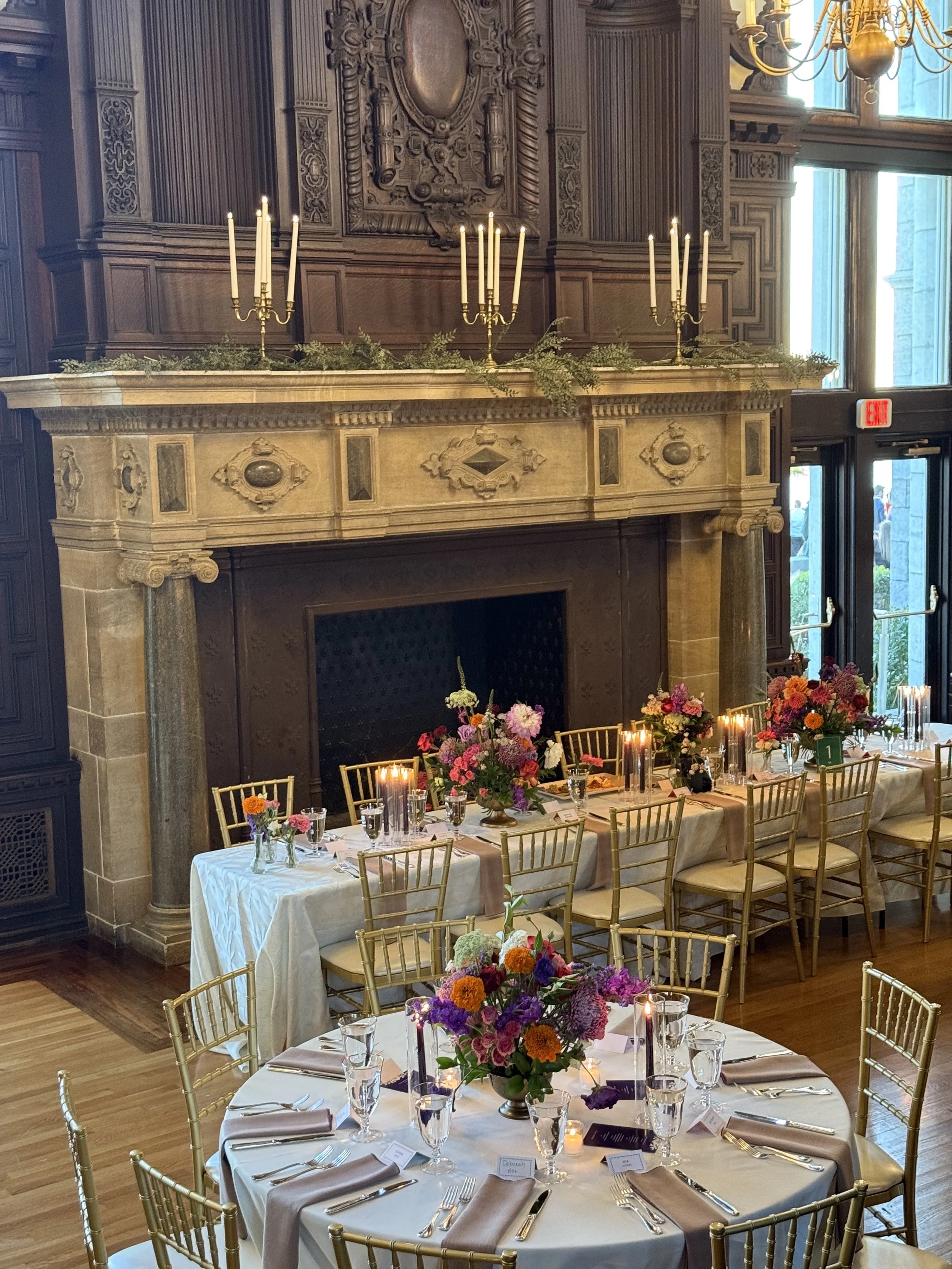The Branford House great room with a large stone fireplace, ornate wood paneling, and a chandelier. Tables with floral centerpieces, gold chairs, candles, and place settings is in the foreground.