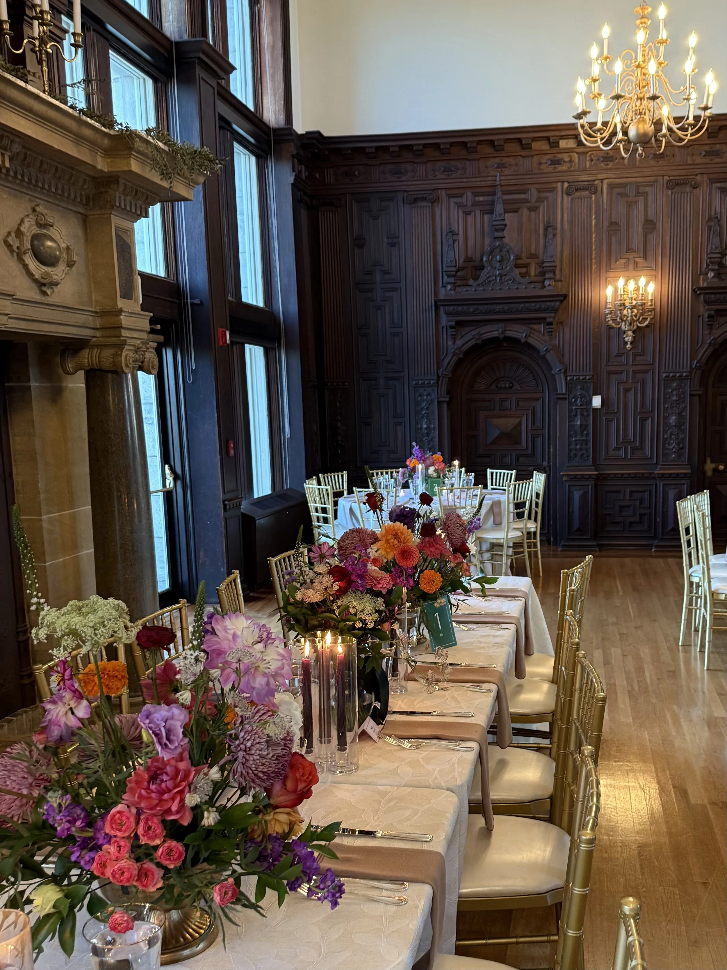 The elegant dining room at Branford House in Groton, CT with wooden paneled walls, large windows, chandelier, ornamental fireplace, and tables decorated with colorful flower arrangements, set for a Clare and Sabrina's wedding