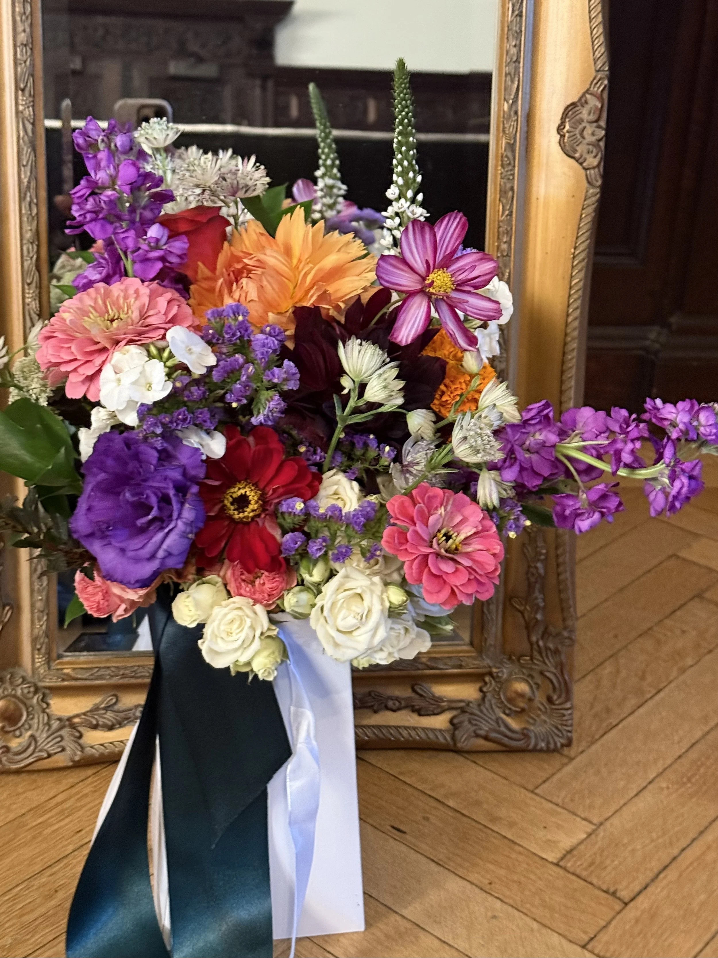 A colorful bouquet of various flowers in a black and white paper wrapper, placed in front of a mirror with a wooden frame on a wooden floor.