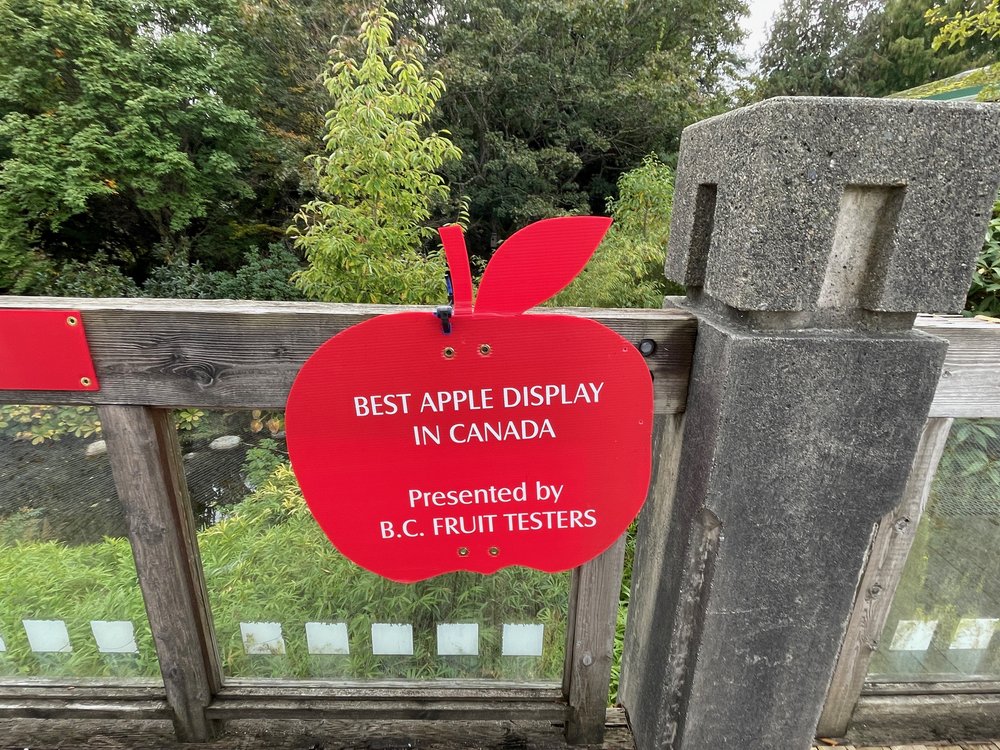  A red apple-shaped sign on a fence, it says “Best Apple Display in Canada, presented by BC Fruit Testers” 