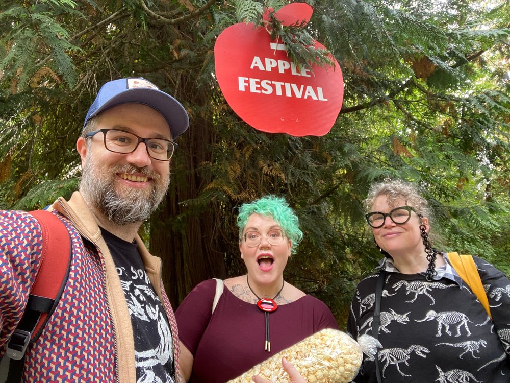  Martin, Hannah and Helen mug for the camera underneath a red apple-shaped Apple Festival sign that is suspended from a tree branch. Hannah holds a long bag of popcorn 
