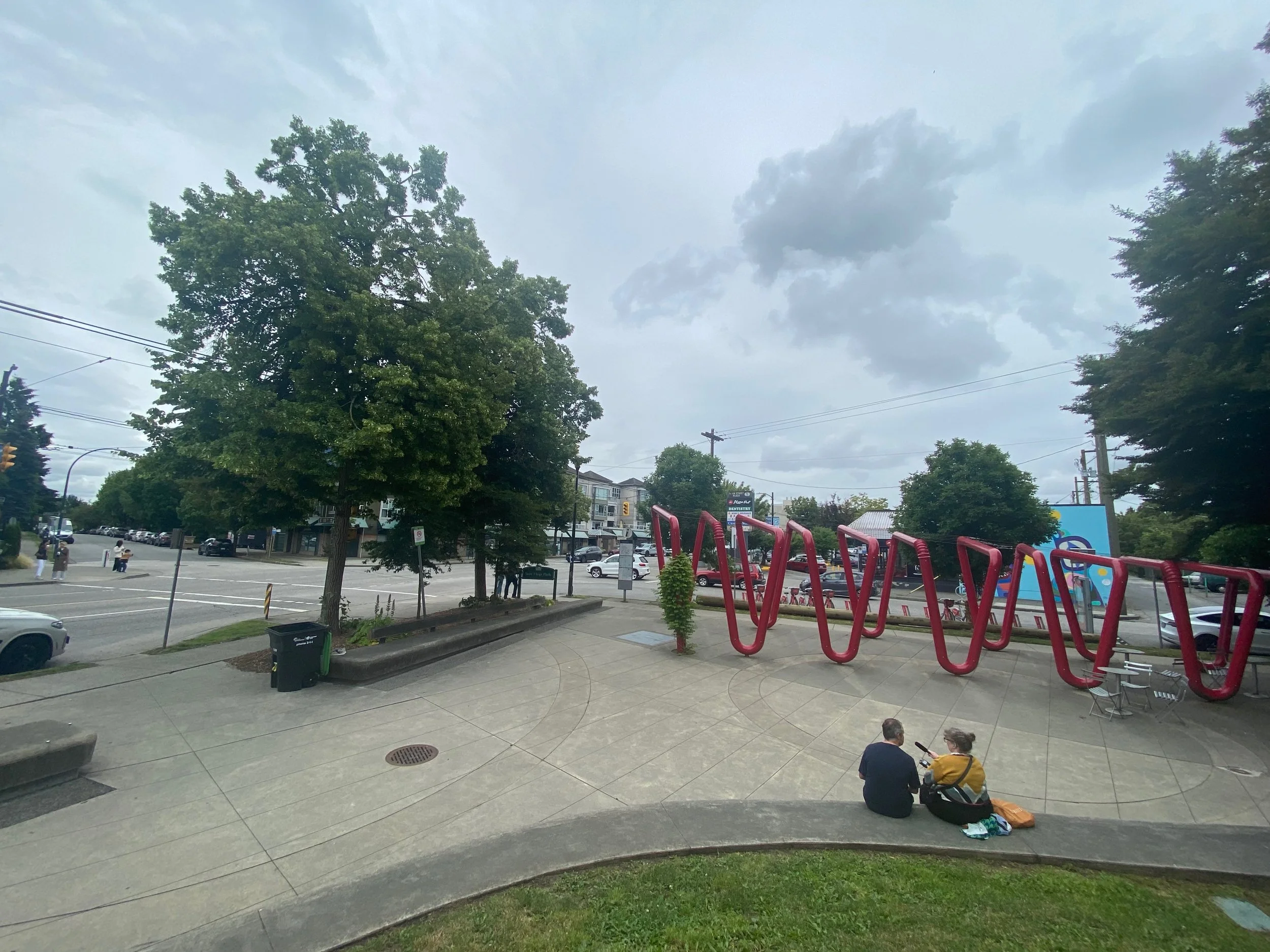   Sun Hop Park:  the view from the nobby hump/hill, of Justin and Helen sitting on the low wall that surrounds it, talking into a microphone. Beyond them is the installation of giant red bendy straws. 