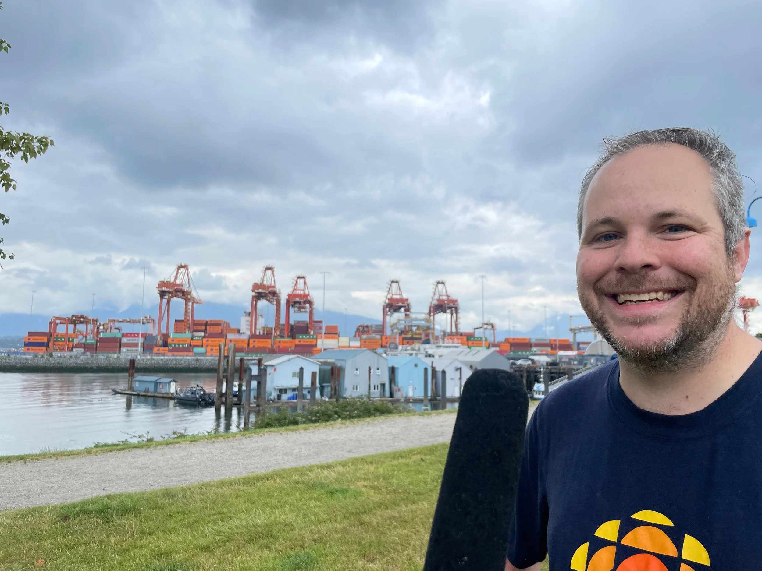   C.R.A.B. Park at Portside:  Justin McElroy stands in the rainy park while Helen waves a microphone in his face. Behind Justin is the port, with large cranes and stacks of shipping containers. 