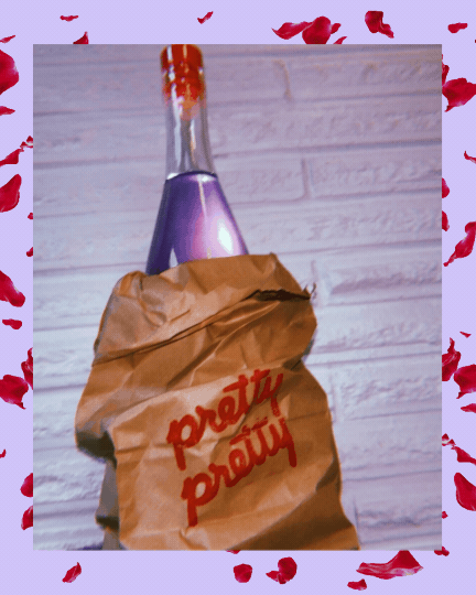 A brown paper bag labeled "pretty prissy" covering a bottle with purple liquid inside, against a light wood wall background, surrounded by scattered red rose petals.