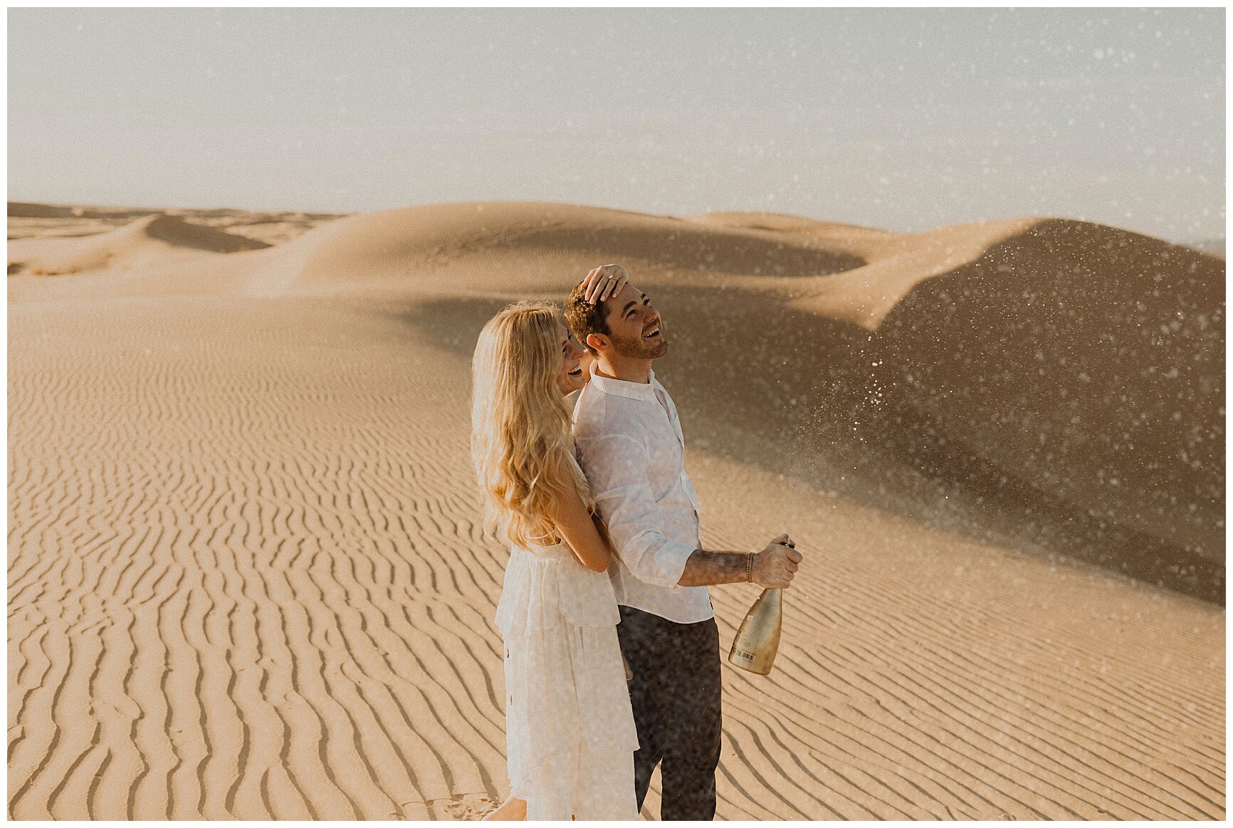  engaged couple popping champagne in the sand during their sand dune engagement session 