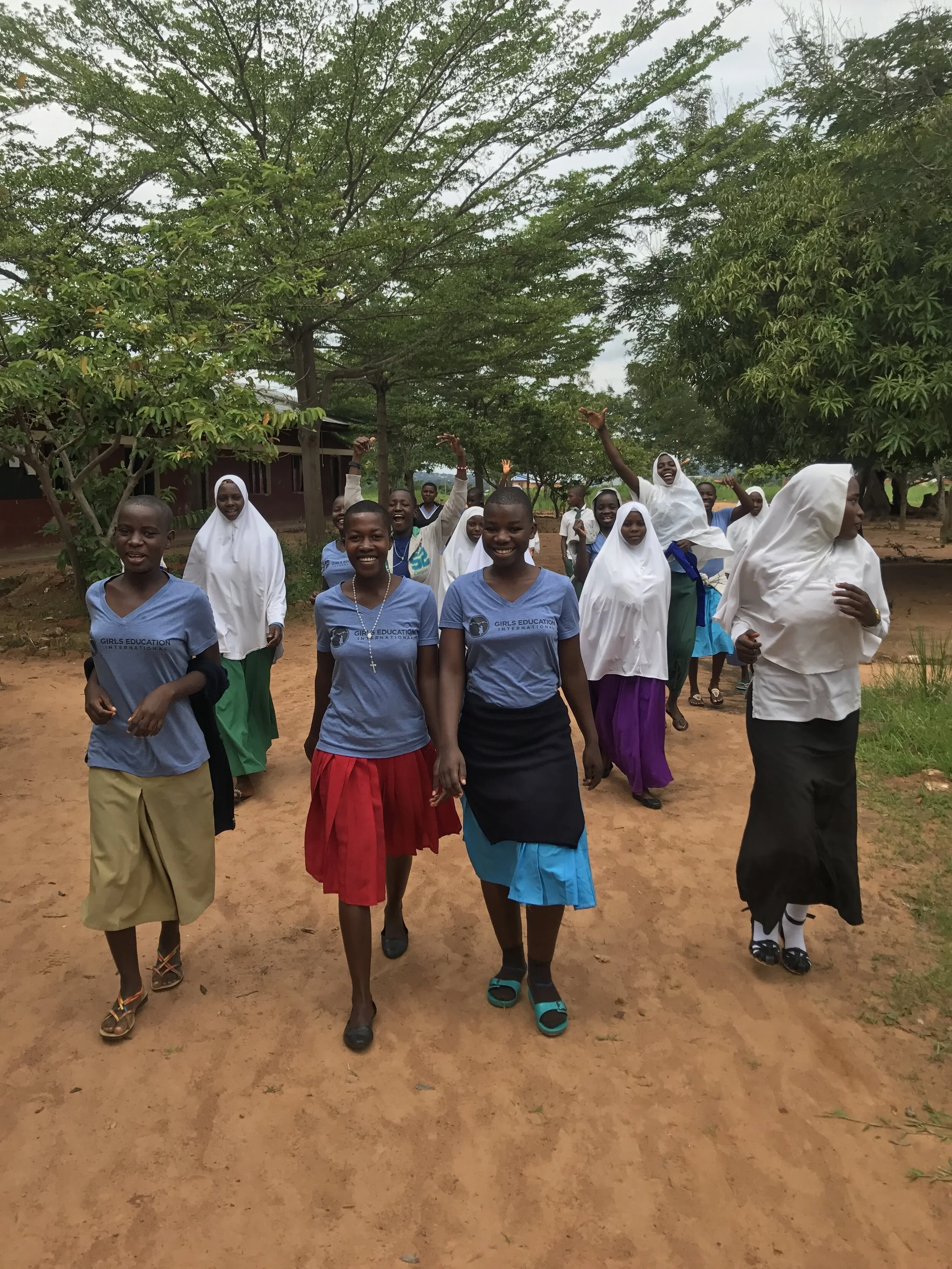 A group of smiling Tanzanian girls walking down a path from school. Some are wearing Girls Education International t-Shirts, some are in hijab.