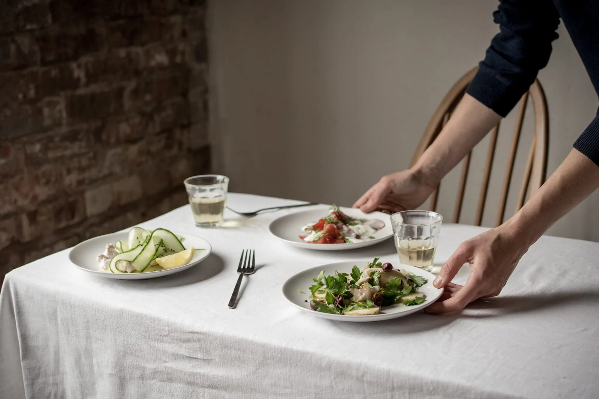 Person setting plates of salad on a dining table with white tablecloth, brick wall in background, and two glasses of white wine.