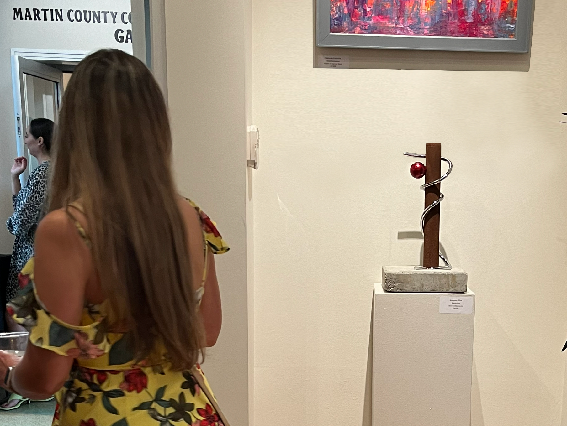 A woman in a yellow dress walking by the sculpture "Paradise," by Norman Silva. The sculpture is made of steel and concrete.