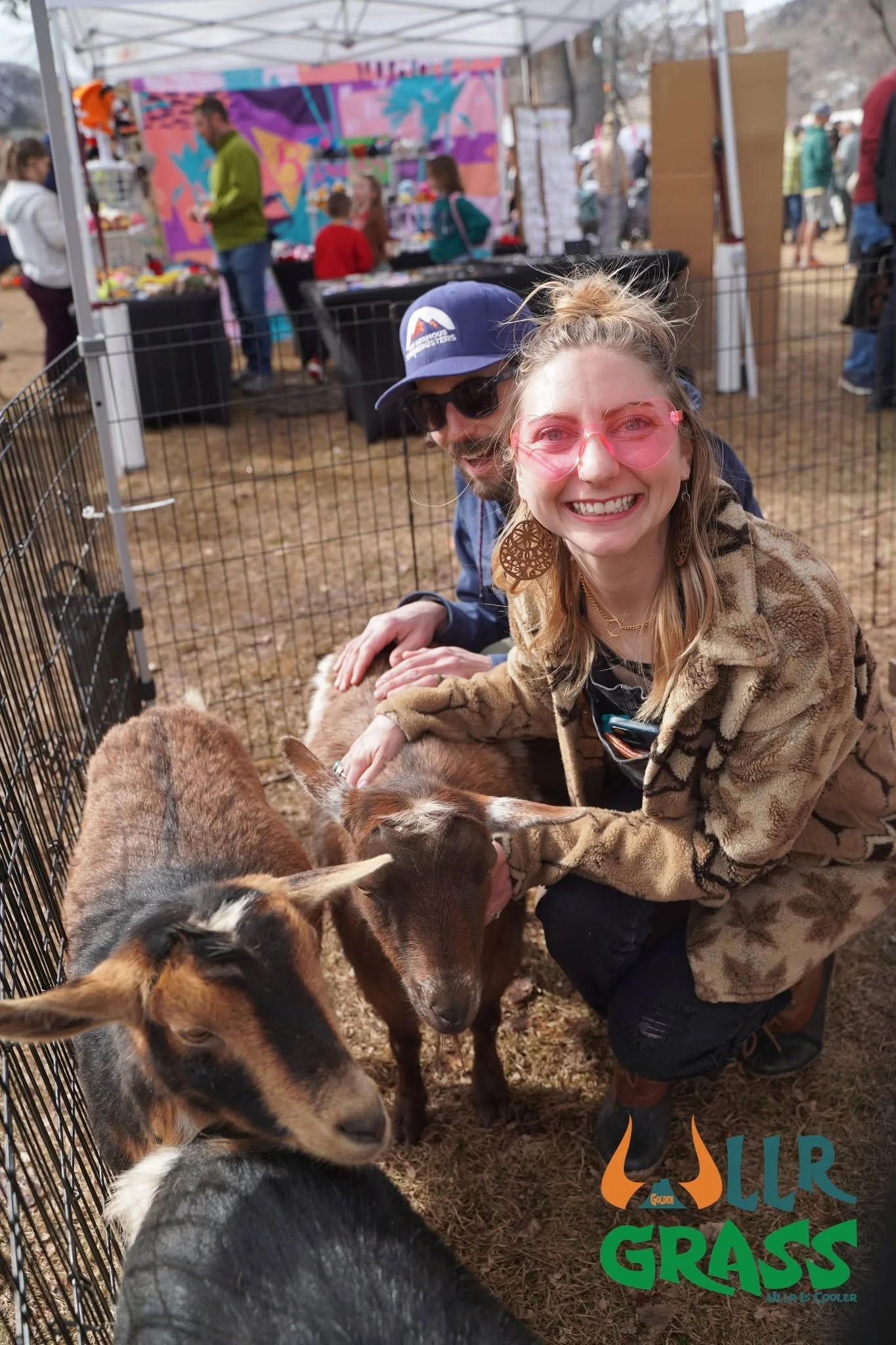 A woman smiling and wearing pink glasses kneels among baby goats inside a fenced area at an outdoor event with booths and people in the background.