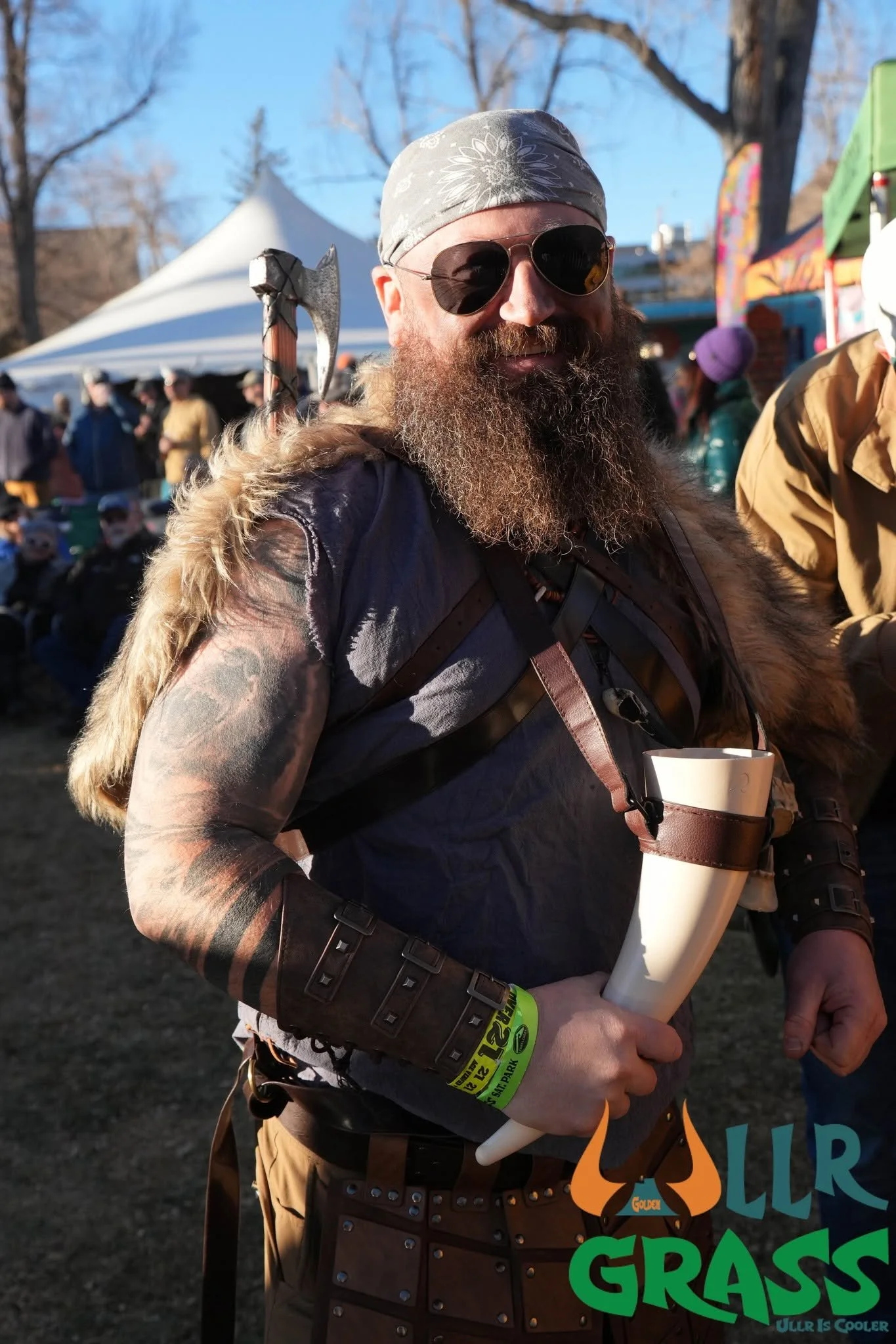 A man with sunglasses, a bandana, and a beard smiling at an outdoor event. He is holding a paper cup and wearing a festival wristband. The background shows tents and other people, with leafless trees and a clear sky.