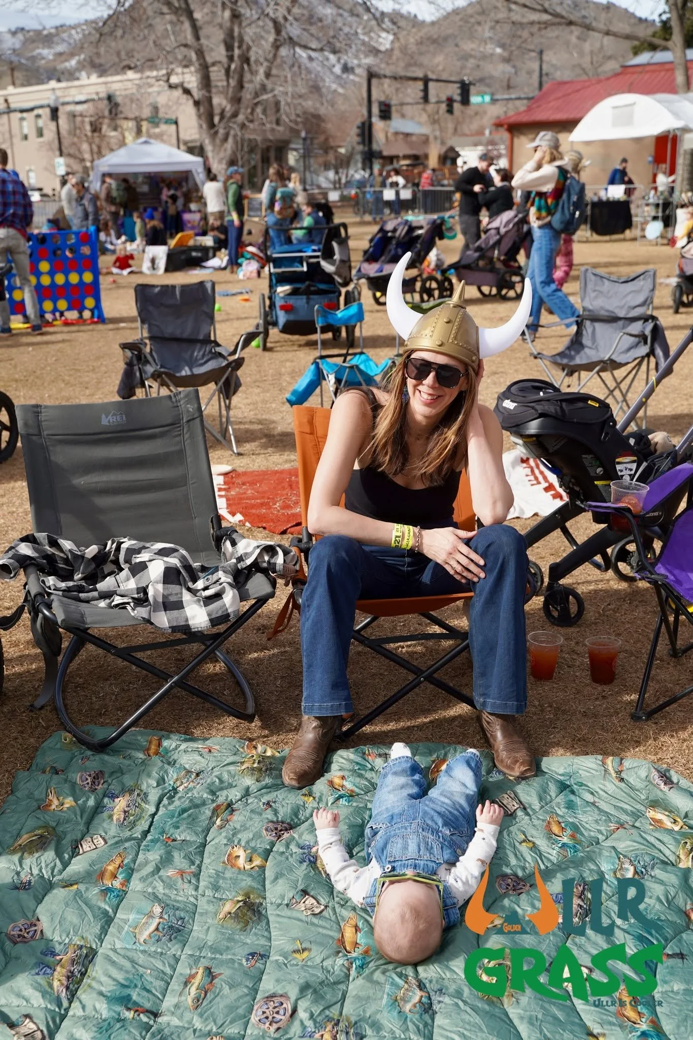 A woman wearing sunglasses and a Viking helmet sits on a camping chair at an outdoor event, smiling at the camera. A young child is lying on a butterfly-patterned blanket in the foreground. Several other chairs, strollers, and people are visible in t