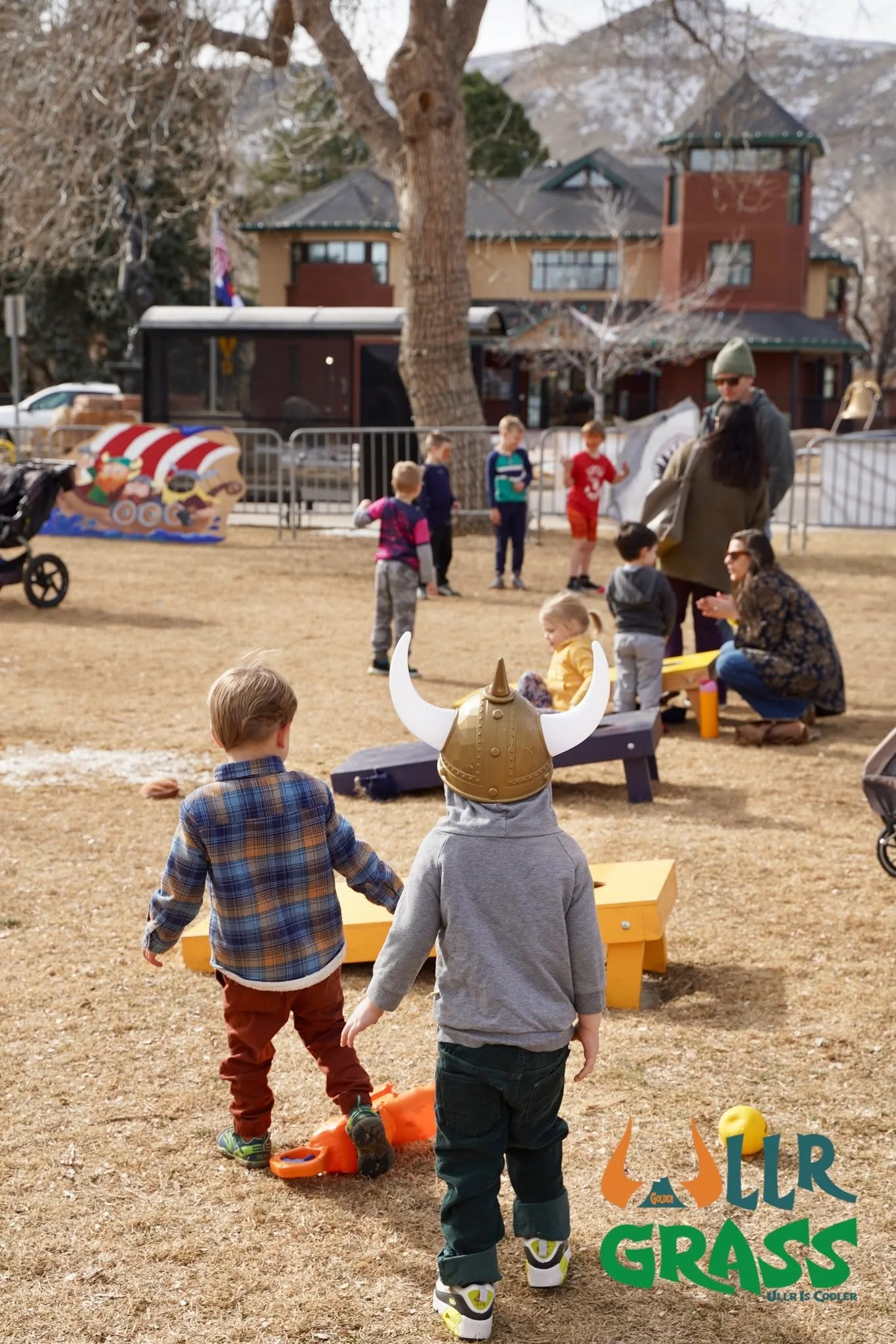 Children playing outdoors on a dry grassy field with adults in the background near a house and trees, some with snow. One child wears a Viking helmet, and others are walking towards a game or activity.