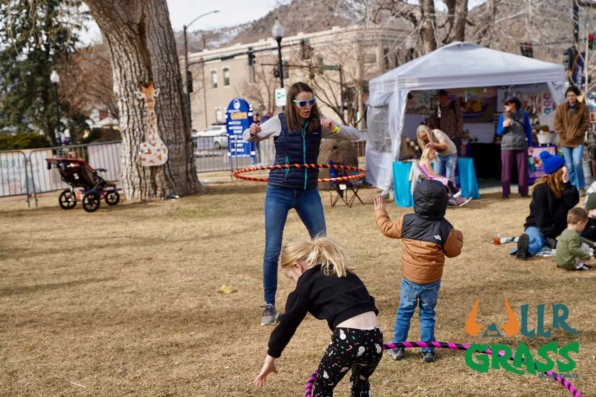 A woman in sunglasses and a navy vest hula hooping at an outdoor event with children and a white tent in the background.
