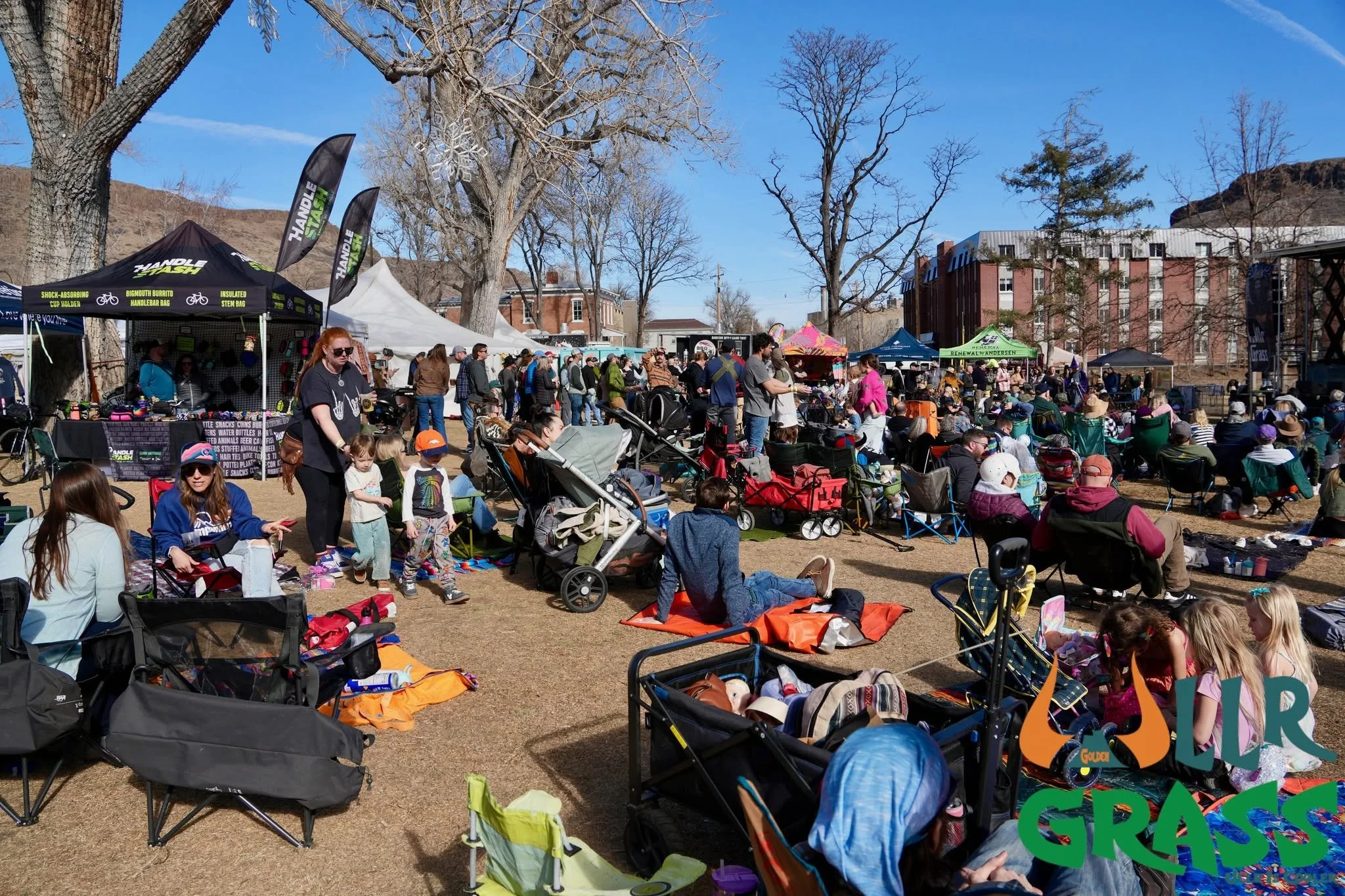 People gather outdoors at a crowded event with tents and booths, some seated on folding chairs and others standing, under clear blue skies with leafless trees and buildings in the background.