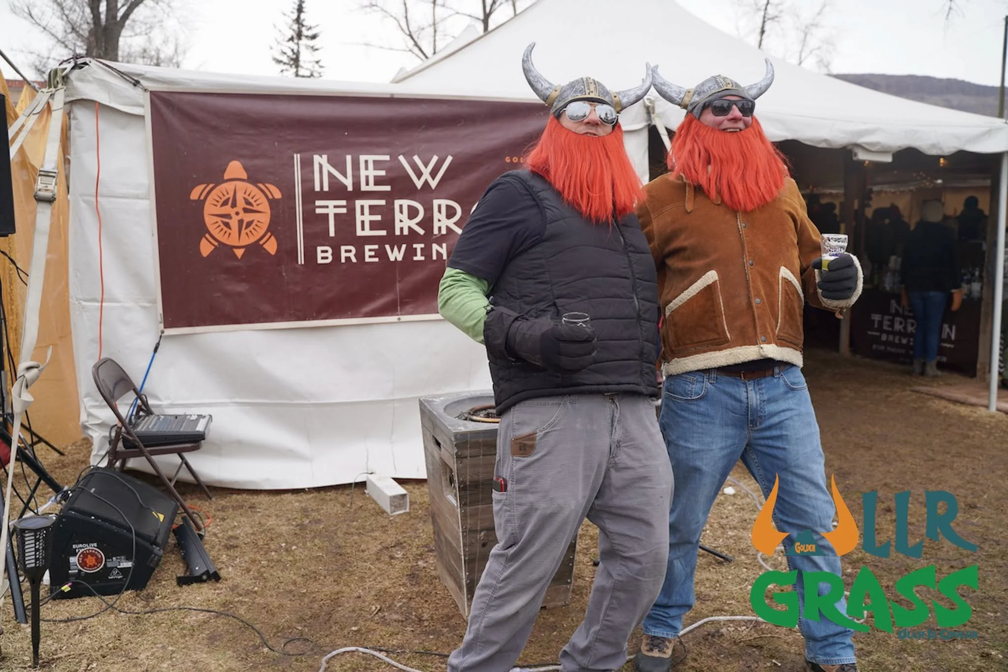 Two men in Viking costumes with orange wigs, horned helmets, sunglasses, and beards standing outdoors at an event, smiling and holding drinks, in front of a sign that reads 'New Terra Brewing'.