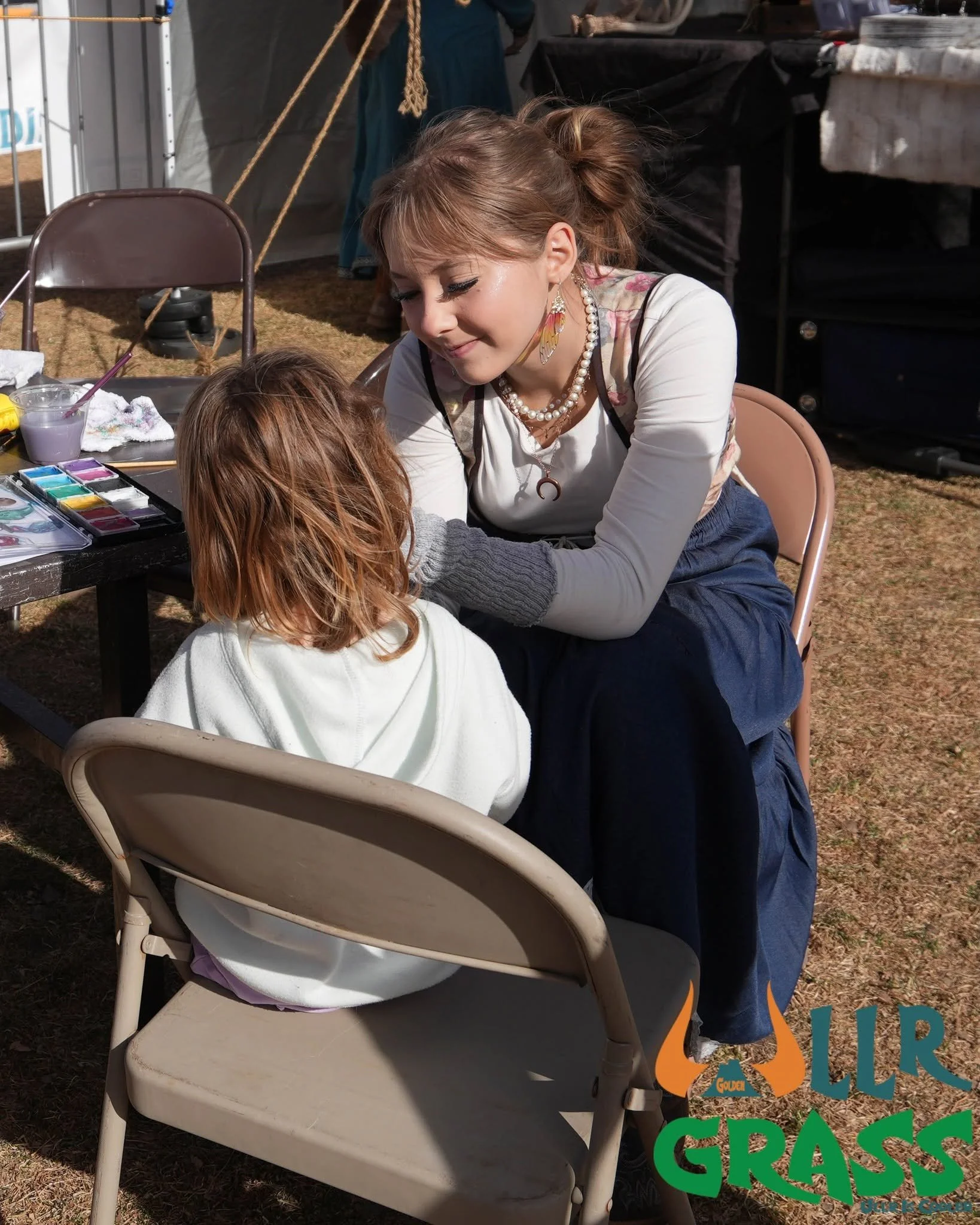 A woman with light brown hair, wearing earrings, a pearl necklace, and a white shirt, is leaning over a young girl with light brown hair, at an outdoor event. The woman is smiling and facing the girl, who is sitting at a table with art supplies, incl