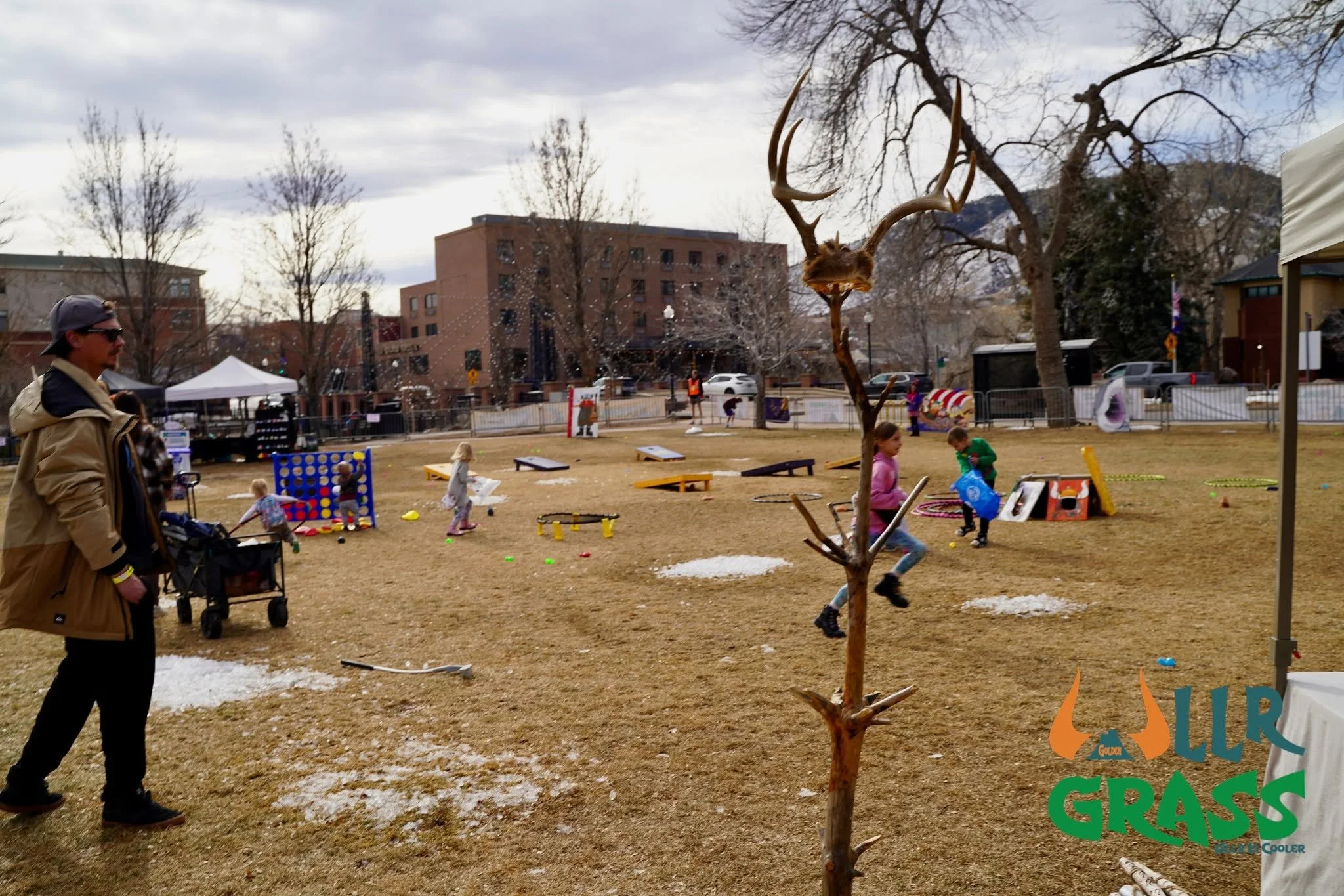 A park with children playing, adults supervising, tents, and trees, with buildings in the background on a cloudy day.