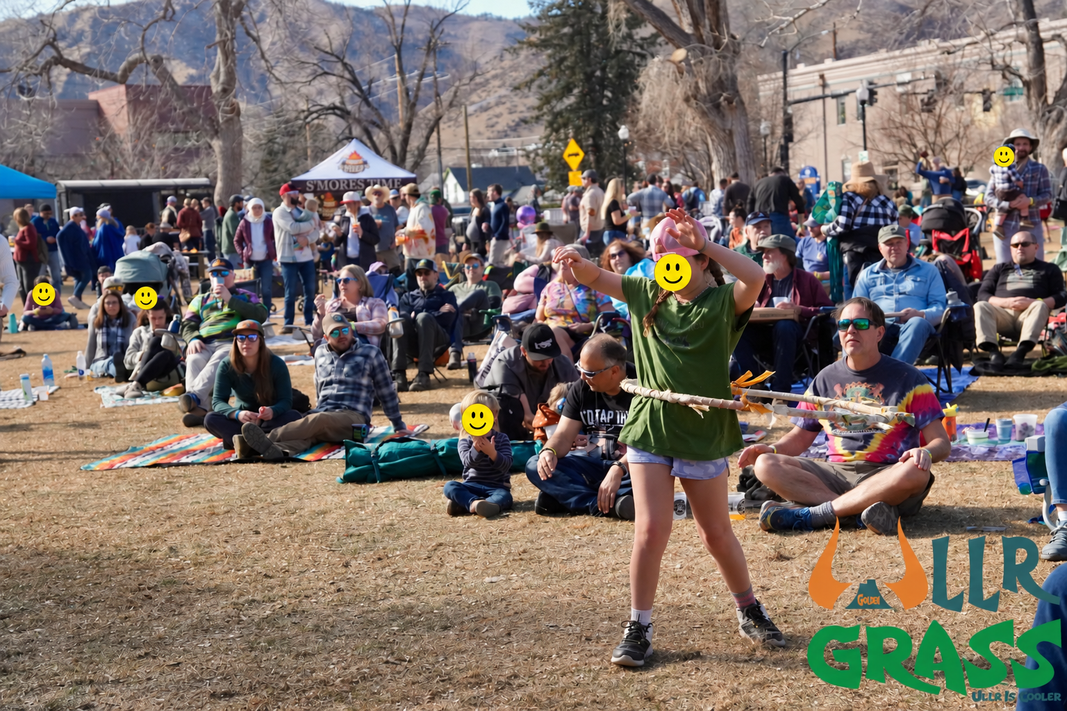 A group of people attending an outdoor event, sitting on the grass and some on blankets, with trees and tents in the background. A girl in a green shirt is dancing or performing in the foreground.