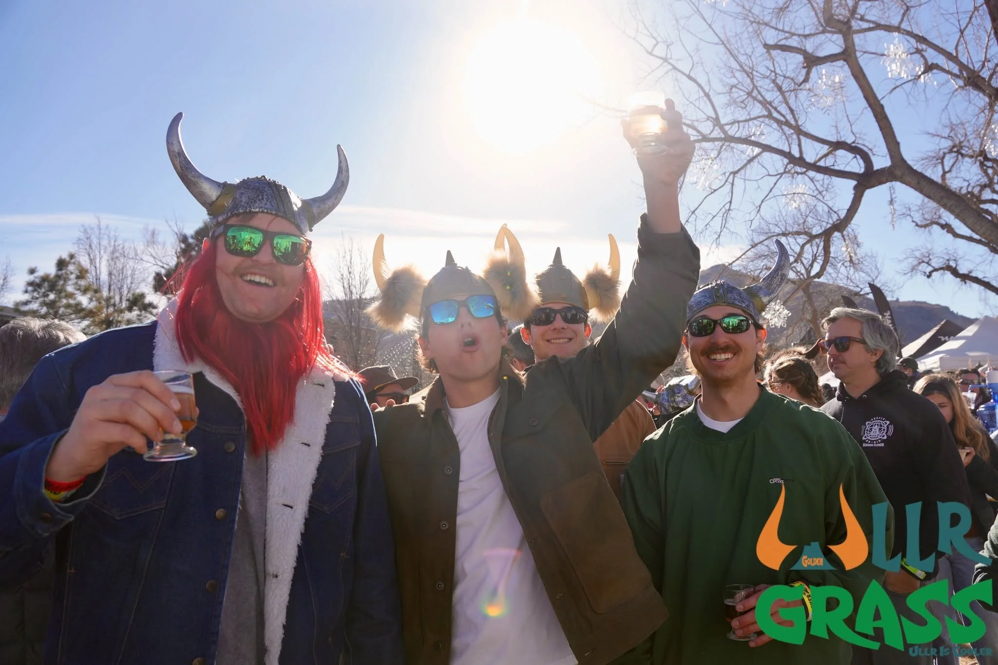Group of people wearing Viking helmets and sunglasses, celebrating outdoors in sunny weather, with mountains and leafless trees in the background, at a St. Patrick's Day event.