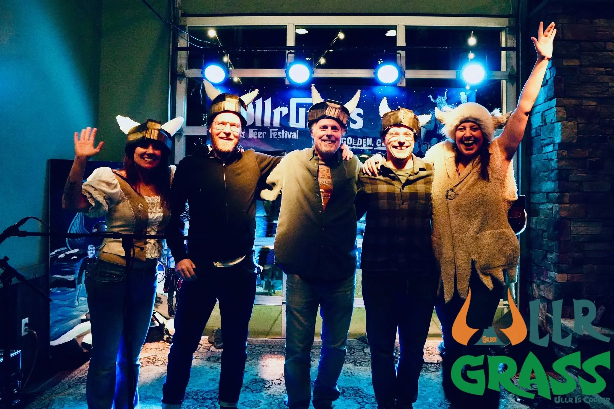 Five people wearing Viking hats with horns and furry hats, standing in front of a stage with bright blue lights and a sign that reads 'All The Girls Beer Festival'. They are smiling and posing for the photo, with two women on either end waving.