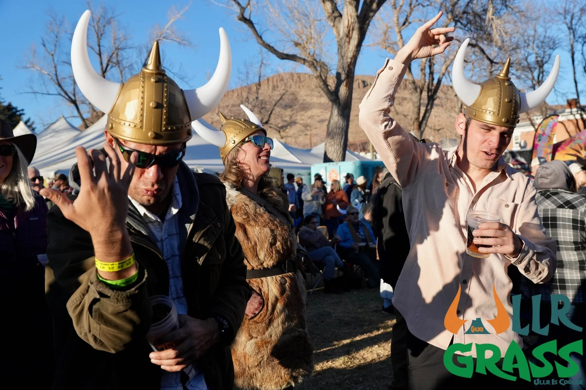 People wearing Viking helmets at an outdoor event, smiling and socializing, with tents in the background and a tree against a clear blue sky.