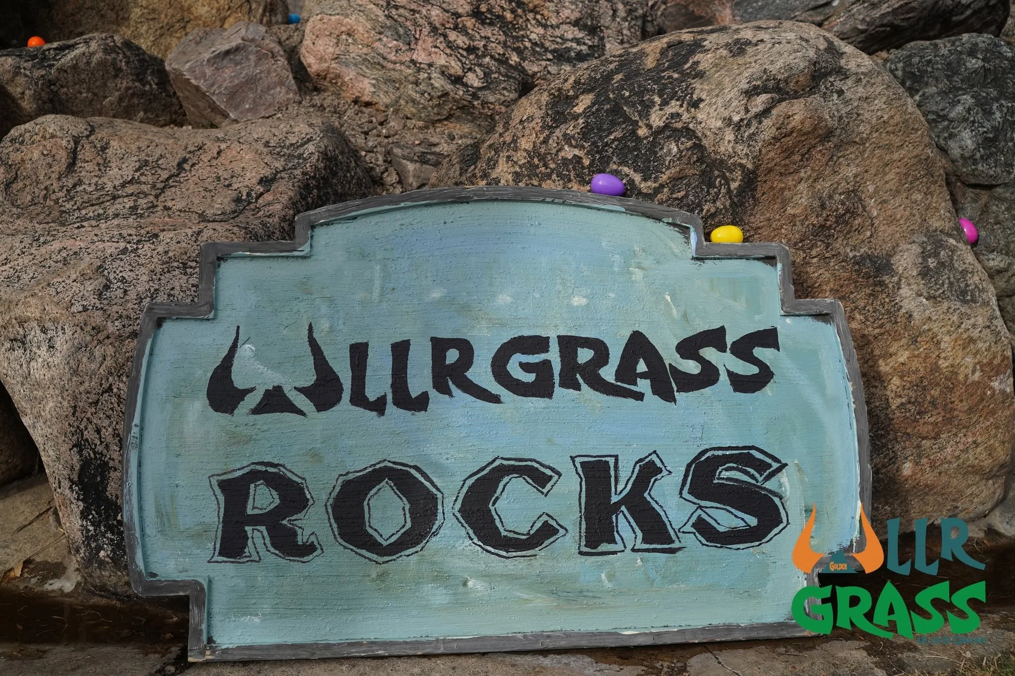A hand-painted wooden sign with the words 'Yes, grass grows' in stylized black letters, placed among large rocks with smaller colorful stones on top.