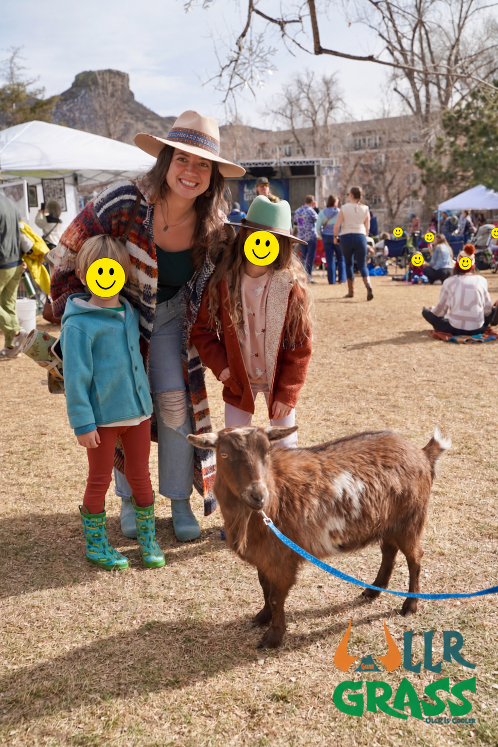 A woman and two children standing outdoors at a farm or outdoor event, with a goat in the foreground and people in the background. The woman is smiling, wearing a large hat and colorful cardigan. The children have smiley face emojis over their faces.