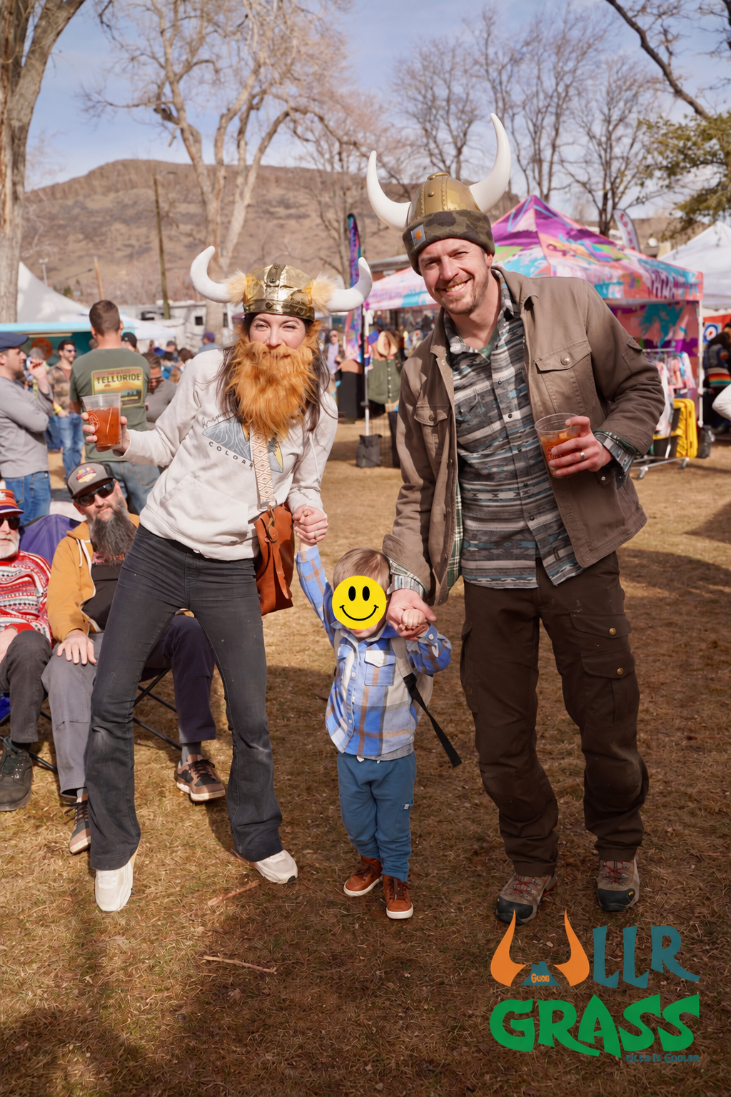 People at an outdoor event, wearing Viking helmets with horns, holding drinks, and smiling, with a colorful tent and trees in the background.