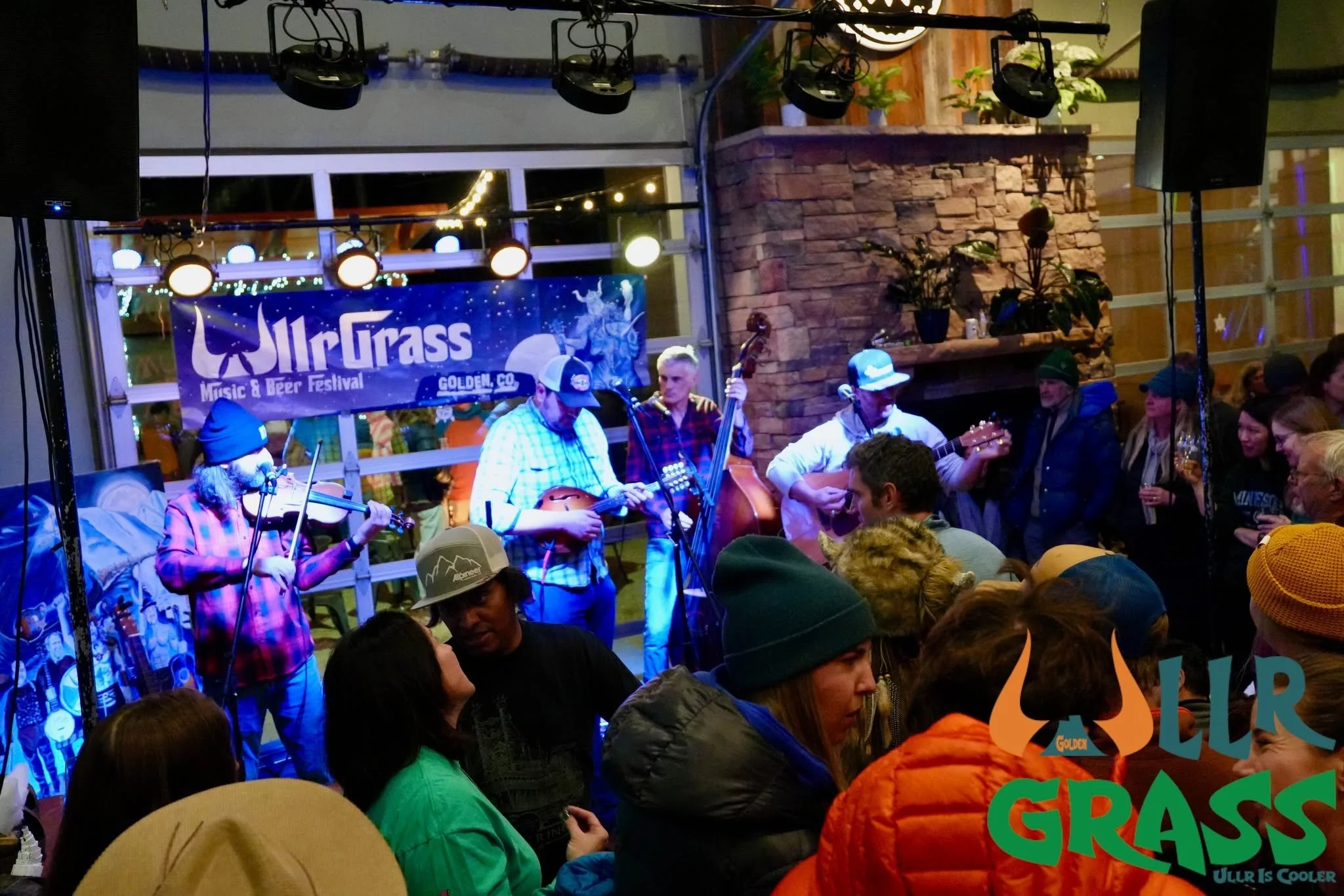 A lively indoor music and beer festival with musicians playing instruments on stage, surrounded by a crowd of festival-goers. The backdrop features a banner for the 'Ulr Grass Music & Beer Festival,' and the setting has warm lighting and decorative plants.
