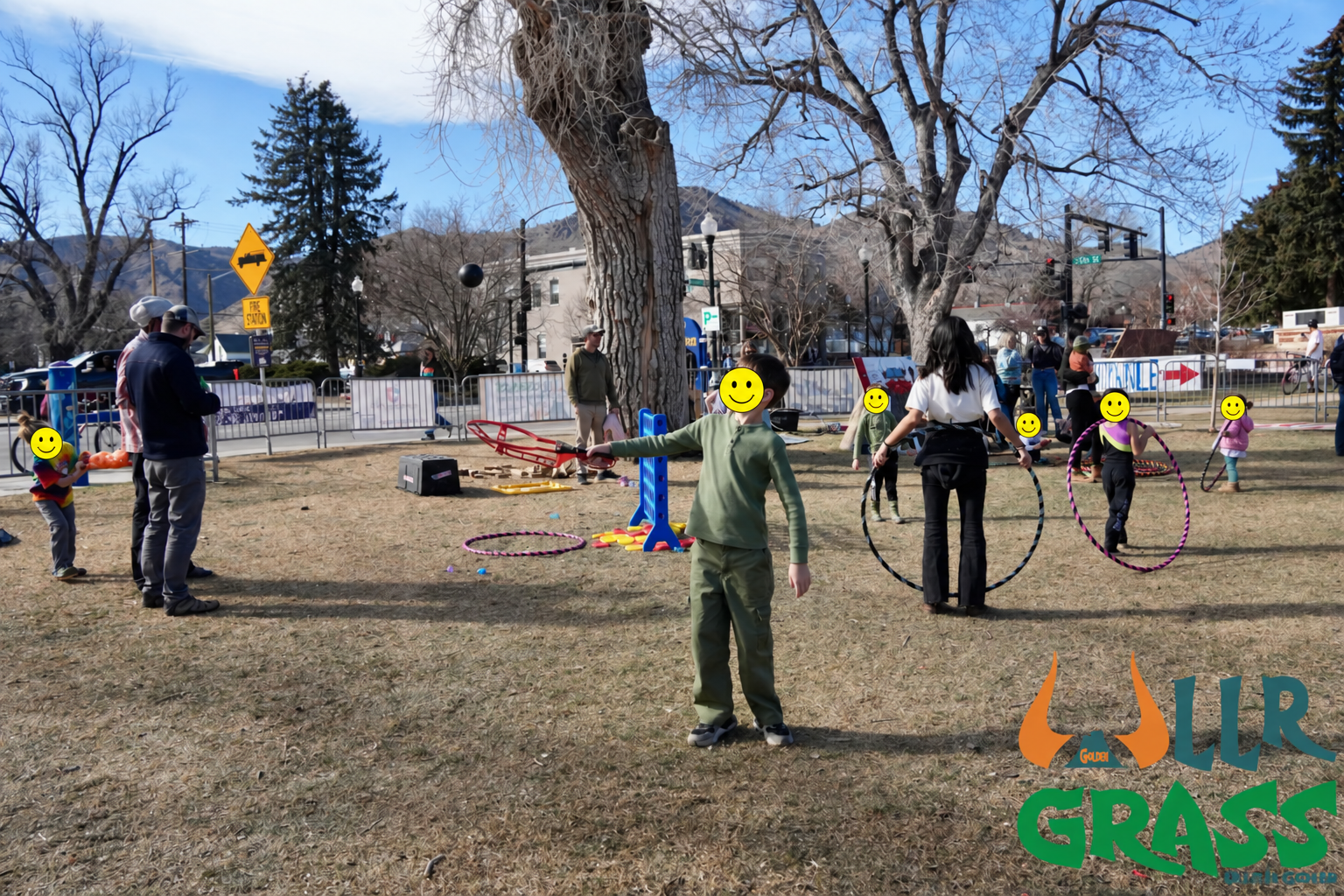 A fenced outdoor event with children and adults, some playing with hula hoops, set in a park with trees and mountains in the background, sunny weather, logo on bottom right reading 'Llr Grass'