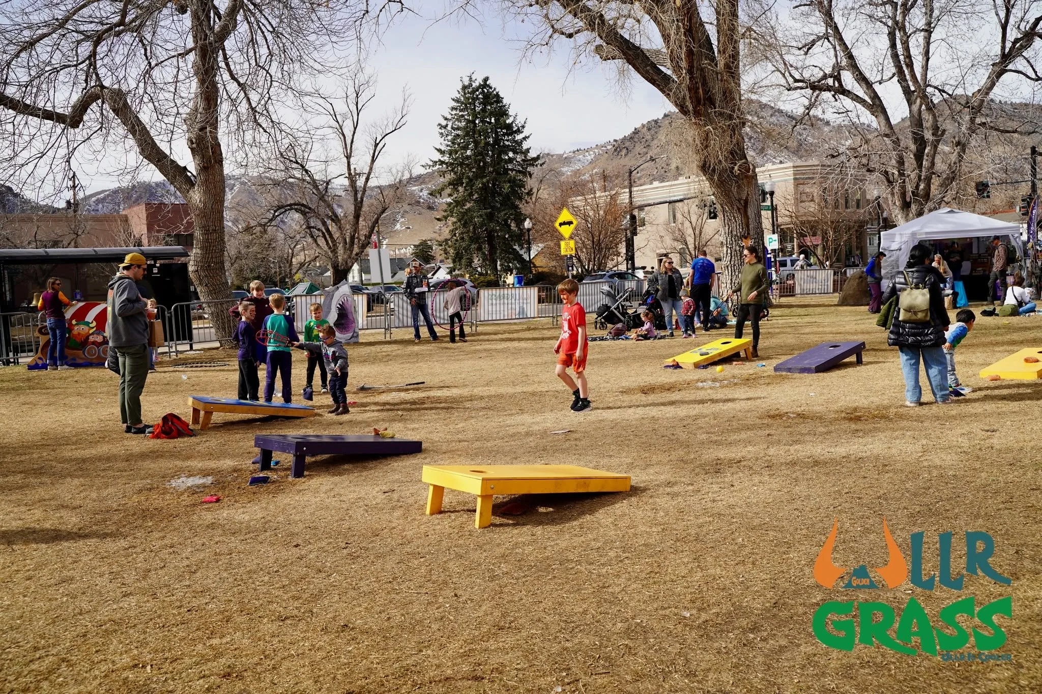 Children and adults playing outdoor games and socializing in a park with leafless trees and mountain backdrop, possibly during early spring or late fall.