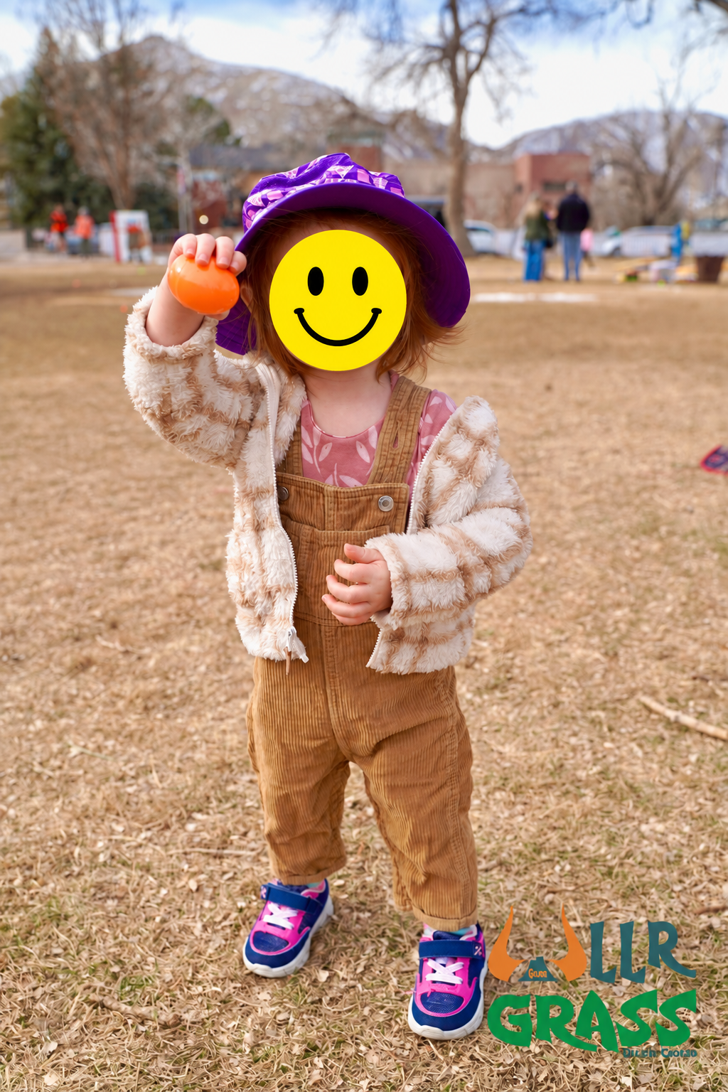 Child wearing a purple hat, fuzzy beige jacket, pink shirt, and brown overalls standing outdoors on grass, holding an orange egg, with a yellow smiley face overlay covering their face, in a park with trees and people in the background.