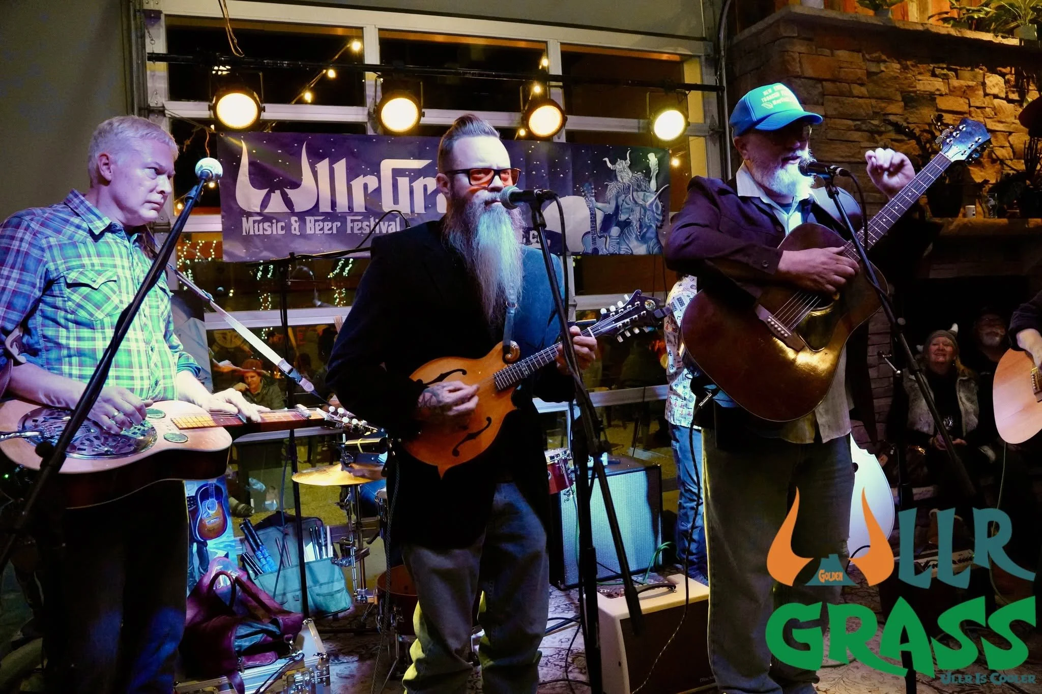 Three male musicians performing on stage at the WallFire Music & Beer Festival, with a colorful banner behind them and warm stage lighting. One is playing a resonator guitar, another a mandolin, and the third an acoustic guitar.