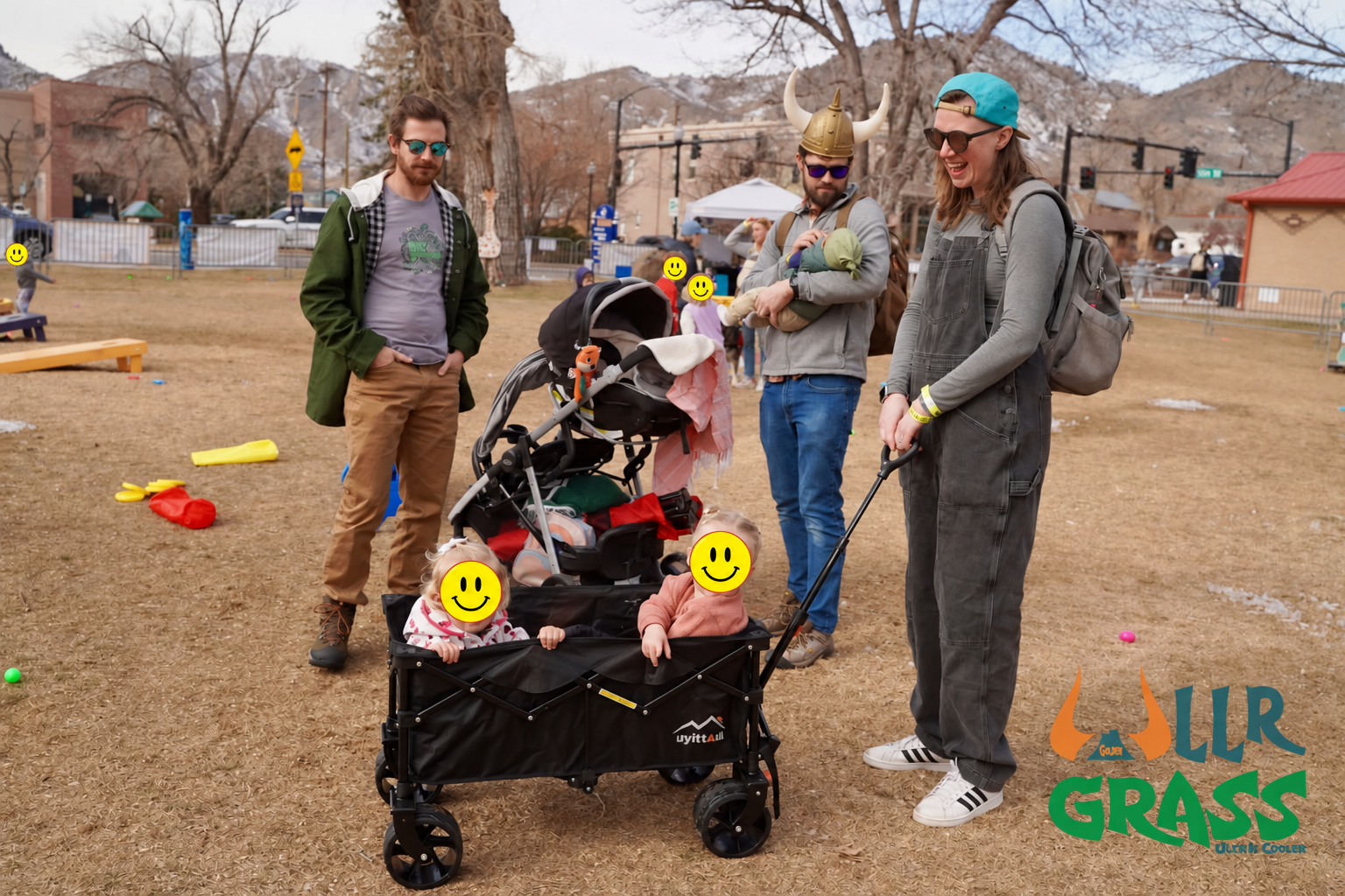 Group of five people, two children, and two babies, standing outdoors in a park with some trees and buildings in the background. One man is wearing a Viking helmet, and a woman is holding a leash. The children are in a stroller, and all faces are cov