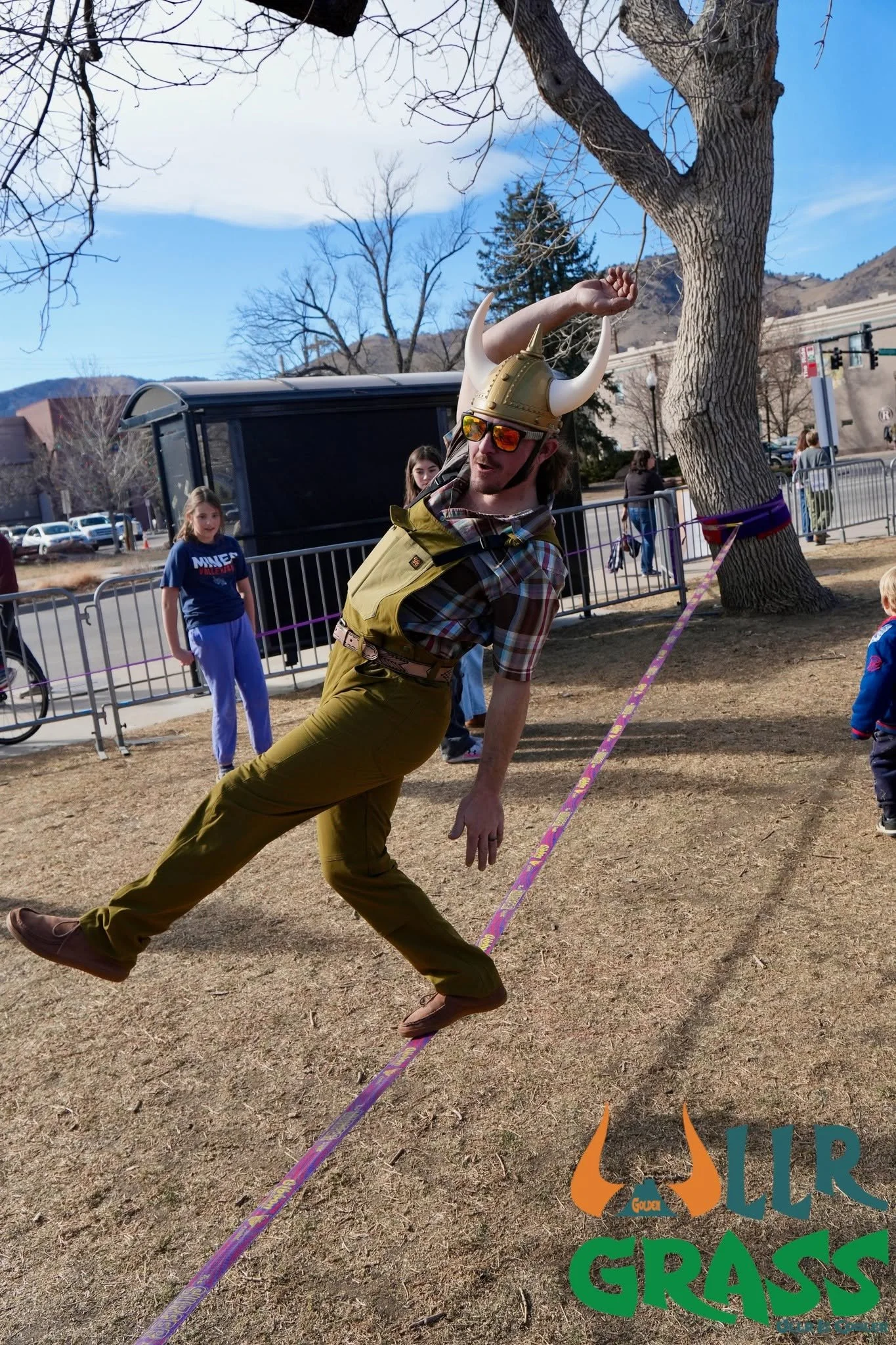 A man wearing a horned Viking helmet, sunglasses, and a plaid shirt posing with a slackline in a park with children and trees in the background.