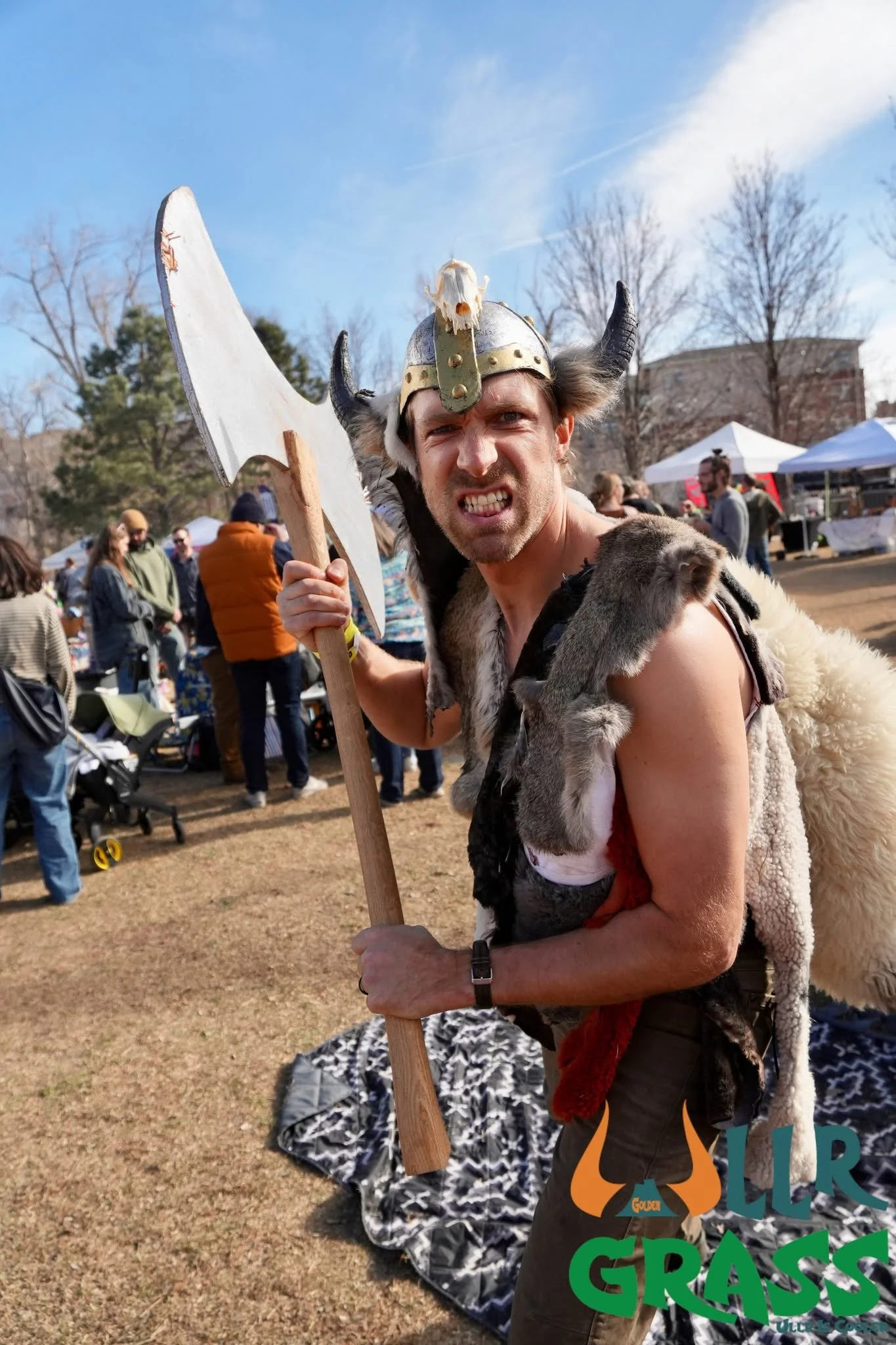 Man dressed as a Viking warrior holding a large axe at an outdoor event. He is wearing a horned helmet with a skull decoration, fur clothing, and has an intense expression. The background shows people, tents, and leafless trees on a clear day.