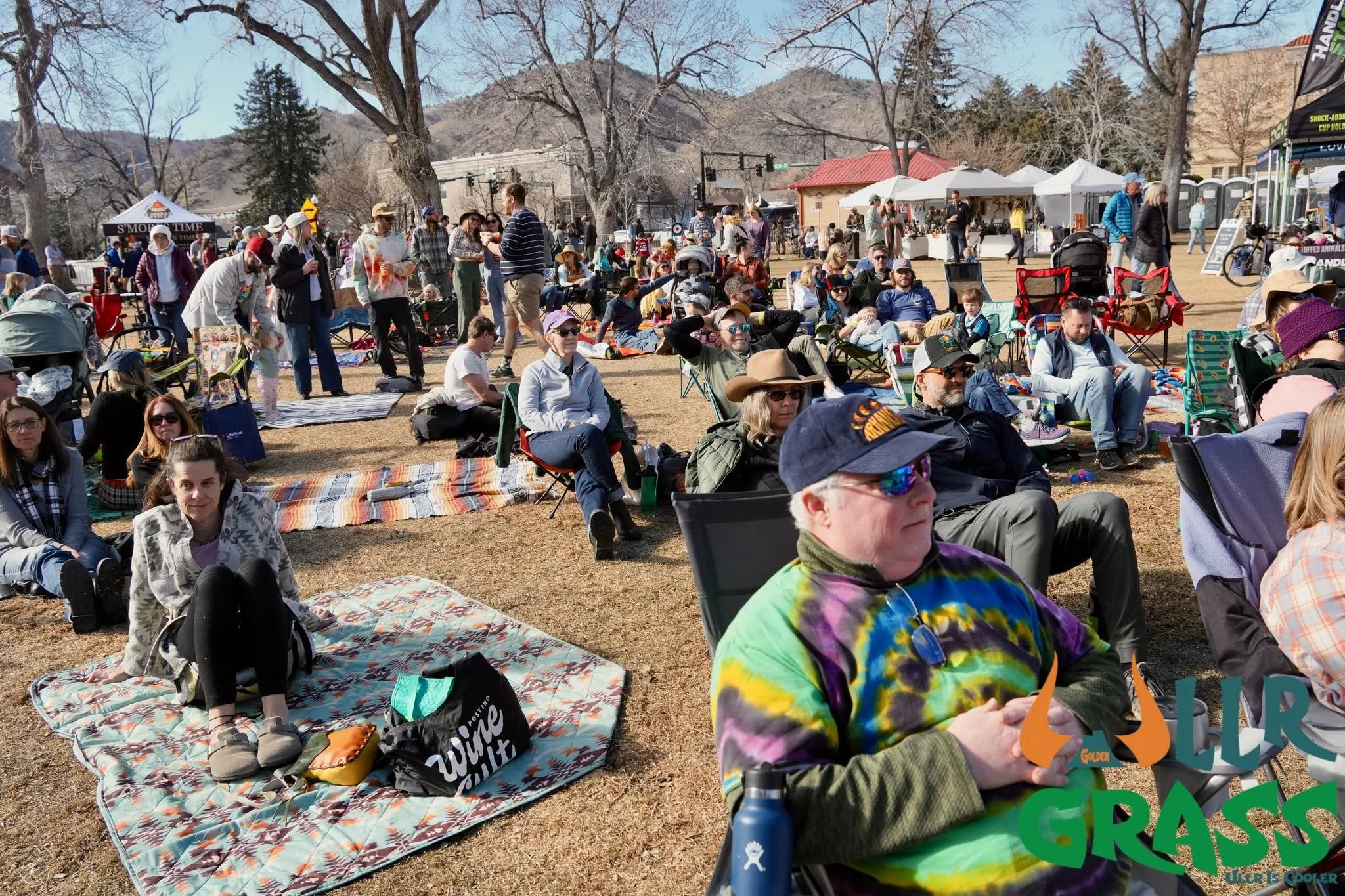 Crowd of people sitting and standing outdoors on a sunny day, some on blankets and chairs, with many tents and booths behind them, mountains in the background.