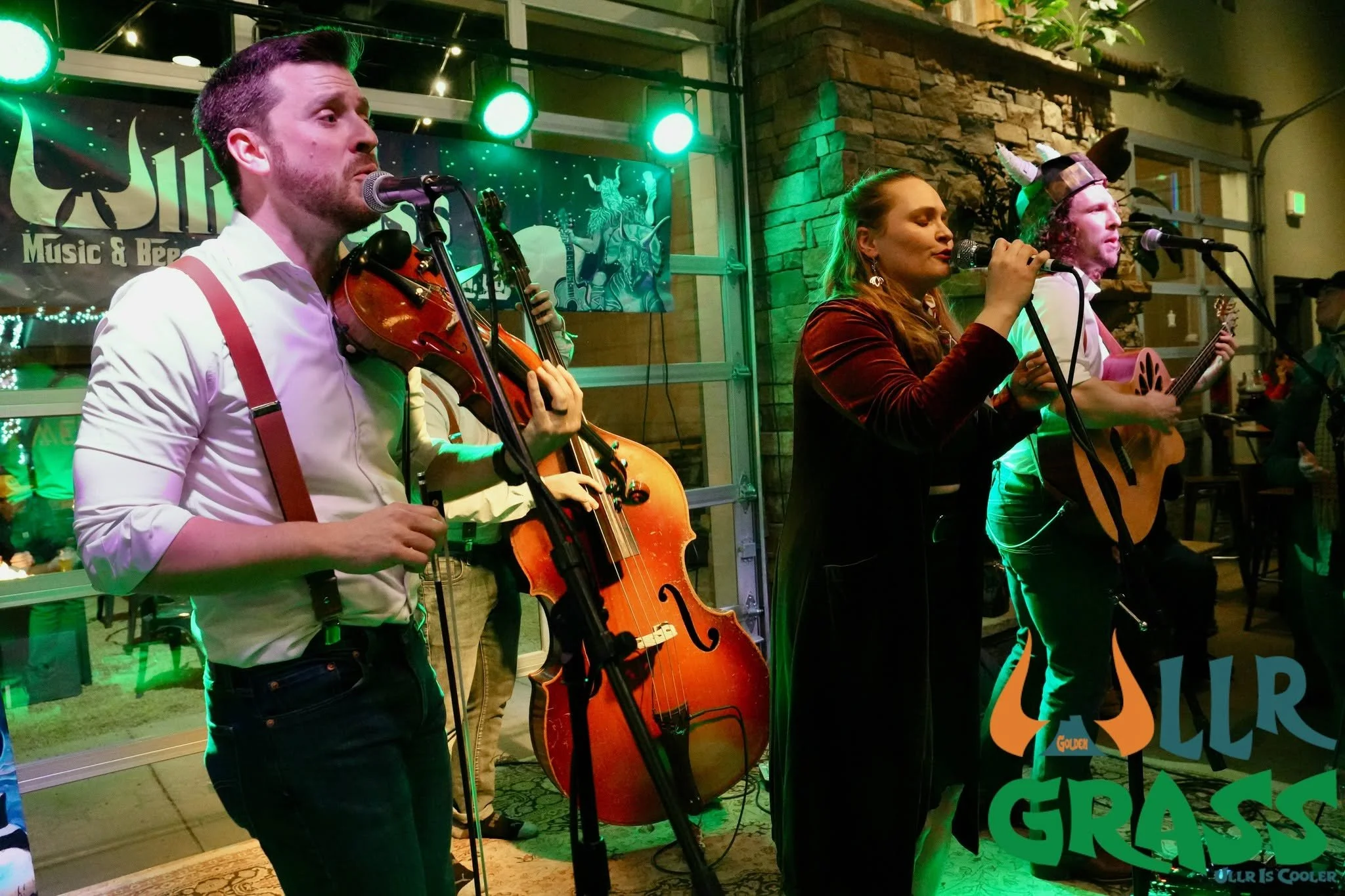 Three musicians perform at a live music event inside a venue decorated with green lighting. The man on the left is playing a violin and singing into a microphone, the woman in the middle is singing into a microphone, and the man on the right is playi