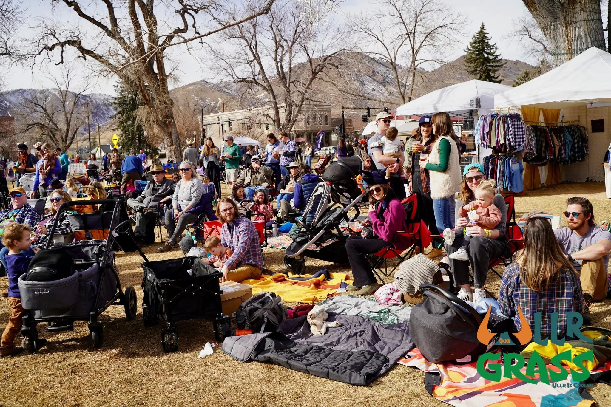 A group of people gathered outdoors in a grassy area, some sitting on chairs or blankets, with several stalls or tents around. The background features leafless trees and mountains, indicating a cool or spring season.